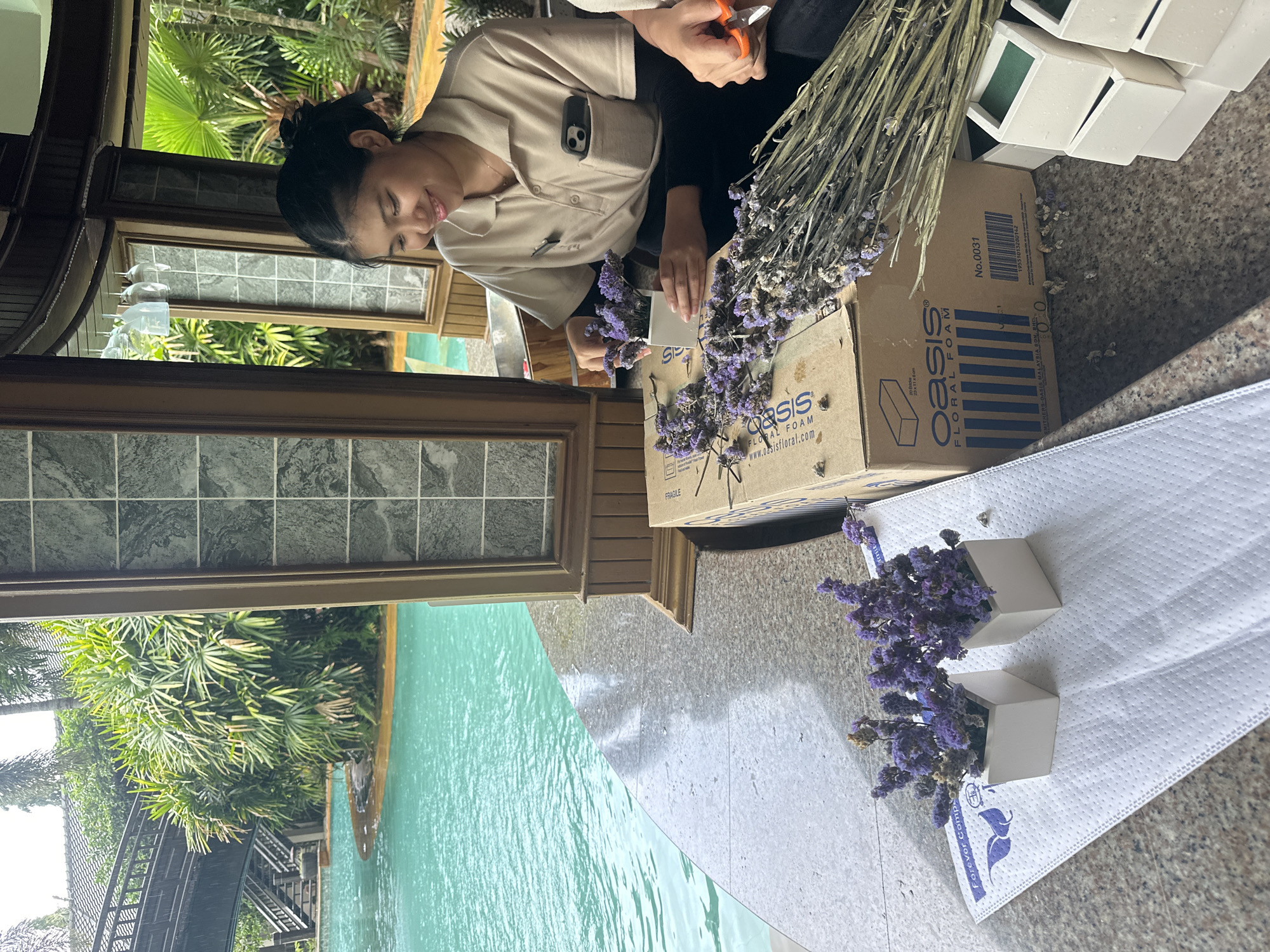 A young woman with dark hair pulled back is smiling as she arranges dried purple flowers in a small white square pot. She is wearing a light brown polo shirt with a black long-sleeved shirt underneath and has a smartphone in her shirt pocket. She is sitting at a counter next to a box labeled "OASIS FLORAL FOAM" and a bundle of dried flowers. In the foreground, there are two white square pots with purple flowers. To the right, another person's hands are visible holding orange-handled scissors and a small white pot. The background shows a bright blue swimming pool, lush green vegetation, and a wooden bridge. The scene appears to be outdoors, possibly at a resort or spa.