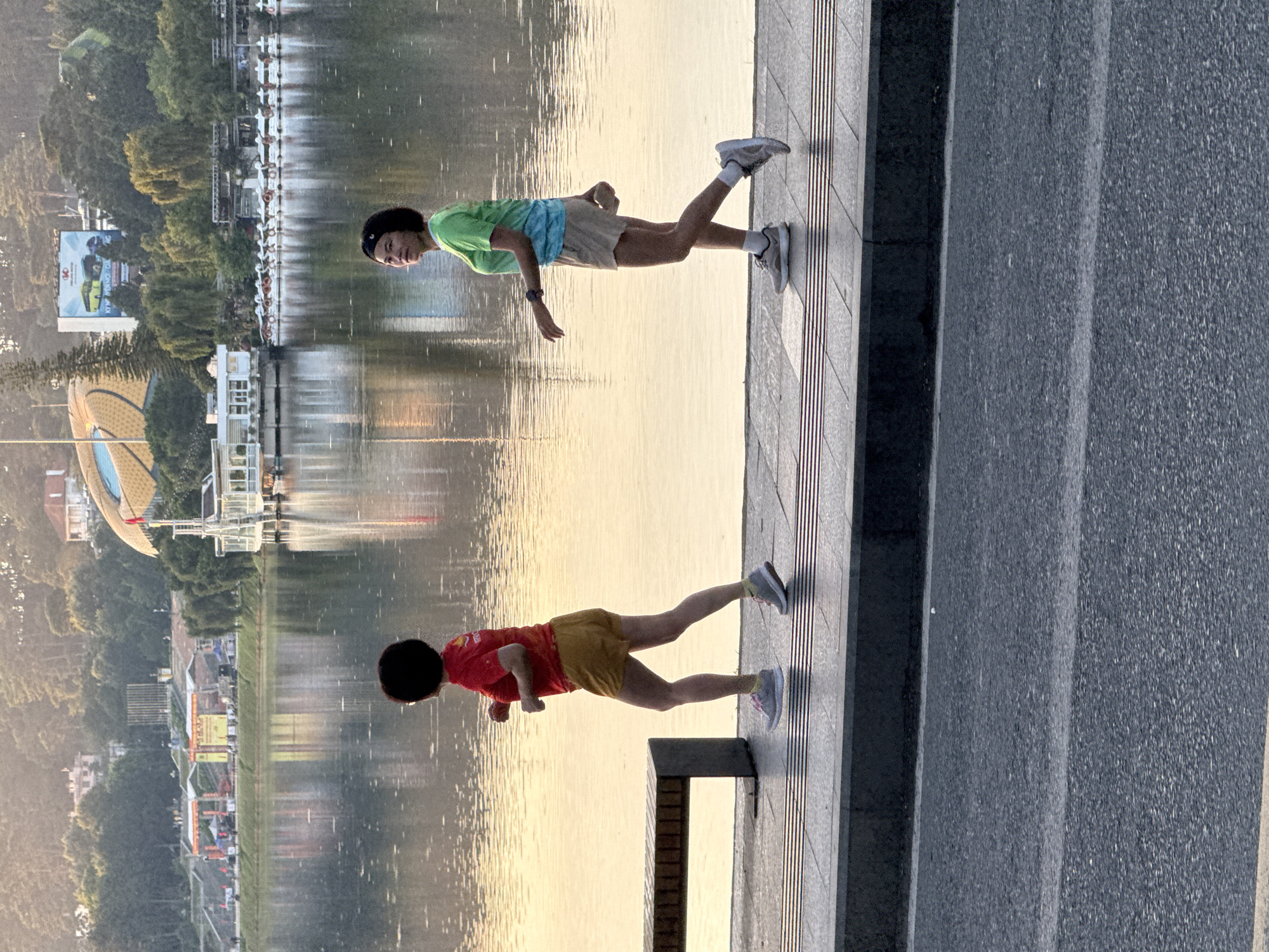 The image depicts Xuan Huong Lake in Da Lat, Vietnam, during either sunrise or sunset, as indicated by the warm, golden light reflecting on the water. Two women are jogging on the paved walkway alongside the lake. The woman on the left wears a red t-shirt and yellow shorts, while the woman on the right is in a green and blue tie-dye t-shirt and beige shorts. A dark wooden bench is visible to the left of the woman in red. In the background, across the lake, are trees, buildings, and a large, distinctive structure with a curved, golden roof. A line of white boats is also moored along the far bank. The foreground shows a road with white lane markings.