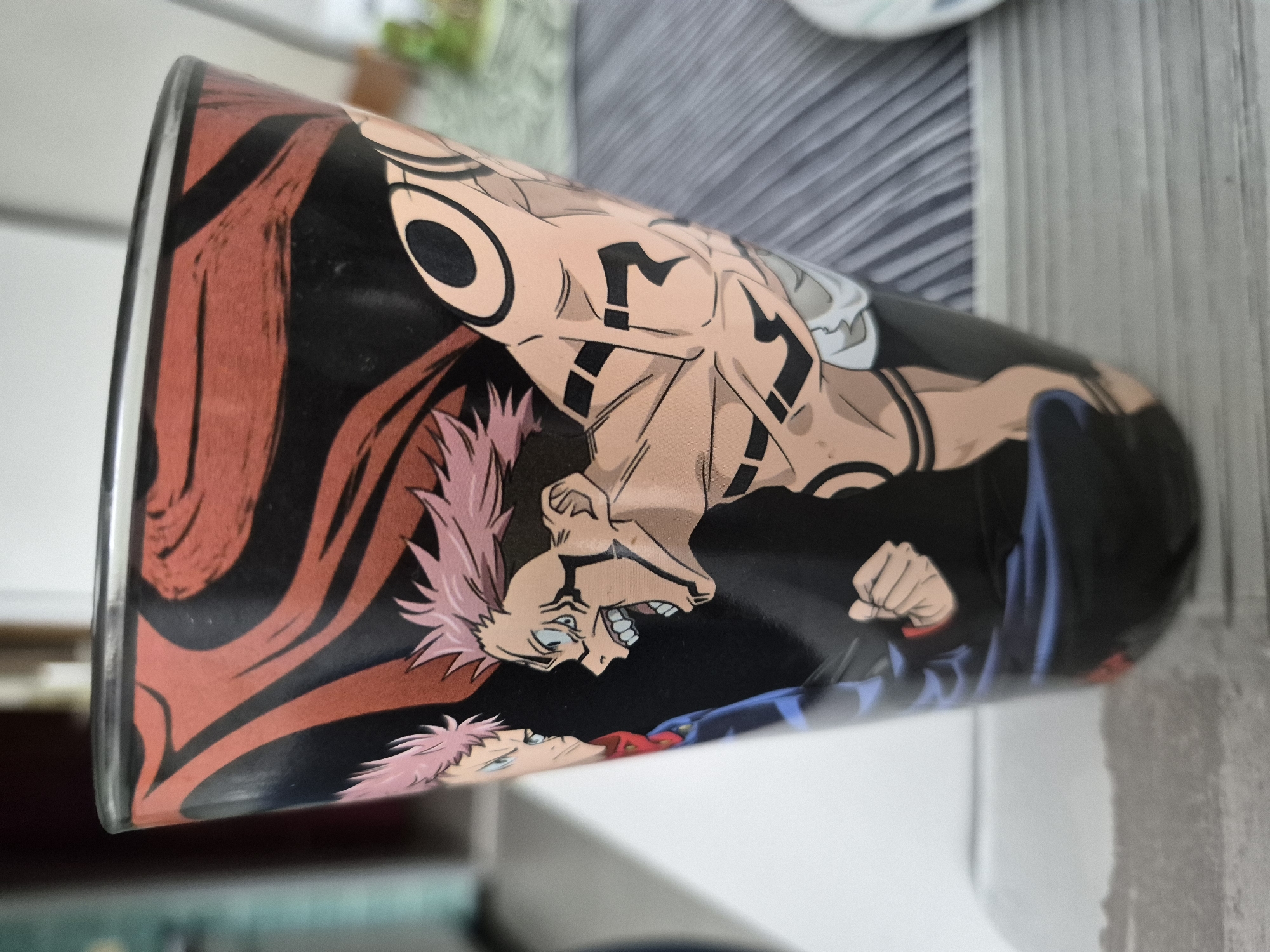 A close-up, slightly angled shot shows the side of a black glass decorated with an anime-style image. The image features two characters with spiky pink hair. The character on the right is depicted with his mouth open in a yell, revealing sharp teeth, and has tribal-like markings on his arms. The character on the left is looking towards the right character, with a more neutral expression, and is wearing a blue uniform with a red collar. The background of the glass's design consists of swirling patterns in red and black. The glass is positioned on a surface that appears to be a table with a striped fabric placemat underneath it, and a lighter, textured surface in the foreground. The background of the environment is blurred, showing hints of furniture and a wall, suggesting an indoor setting.