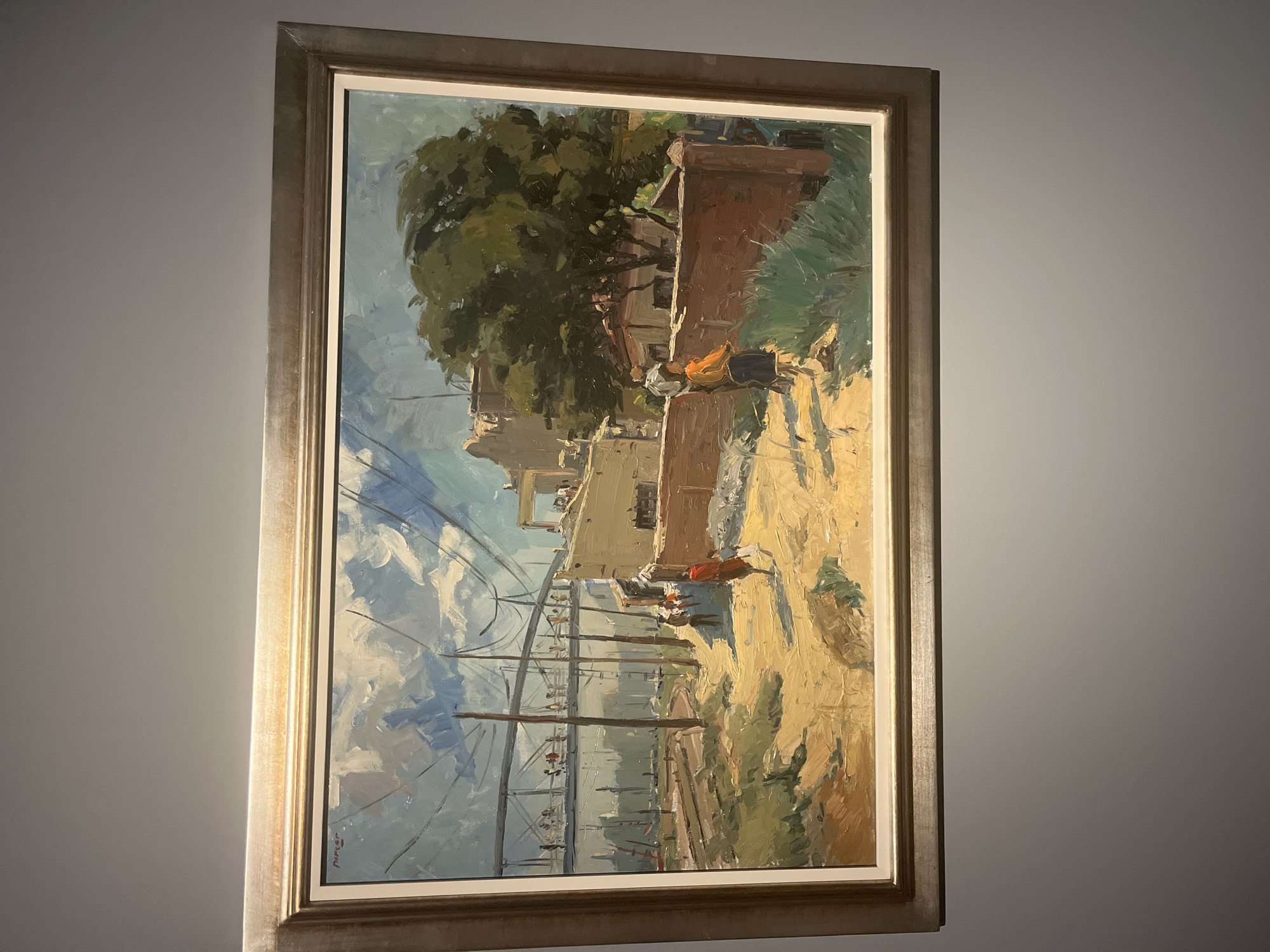 An impressionistic painting, likely oil on canvas, depicts a vibrant street scene or pathway in a sun-drenched, possibly Mediterranean, environment, set in València, Spain. The artwork is presented in a simple, metallic-toned frame.
The scene is dominated by a bright blue sky with dynamic white clouds. In the upper left, a large, lush green tree with dense foliage provides a stark contrast to the light-colored buildings with flat roofs that stretch across the mid-ground. A prominent feature of the painting is a network of tall utility poles and numerous crisscrossing wires that extend across the left and central parts of the canvas, adding a sense of modern infrastructure to the rustic setting.
Several small, loosely rendered figures are visible. In the mid-ground to the right, a figure wearing an orange or red garment walks along a dusty, sunlit path, which forms a significant part of the foreground in shades of yellow, beige, and brown. Closer to the viewer and towards the center-left, a small group of figures appears to be gathered or walking near the base of the utility poles. The ground is unpaved and appears dry and dusty.
The painting conveys a strong sense of bright daylight, possibly late morning or afternoon, through its vivid color palette and the interplay of light and shadow. The brushstrokes are visible and energetic, giving the piece a lively and immediate feel. An artist's signature, possibly "J. Pinedo" or "J. Pantoja," is discernible in the bottom left corner of the painting.