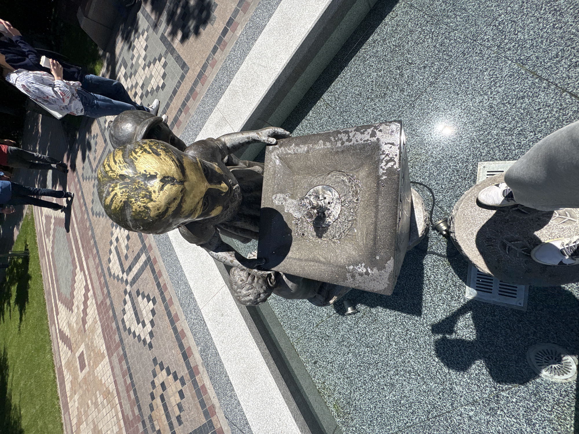 The image depicts a bronze statue of a child positioned beside a drinking fountain in Yerevan, Armenia. The fountain is active, with water visibly flowing. People are standing nearby. The surrounding area includes a patterned brick walkway and grassy areas.