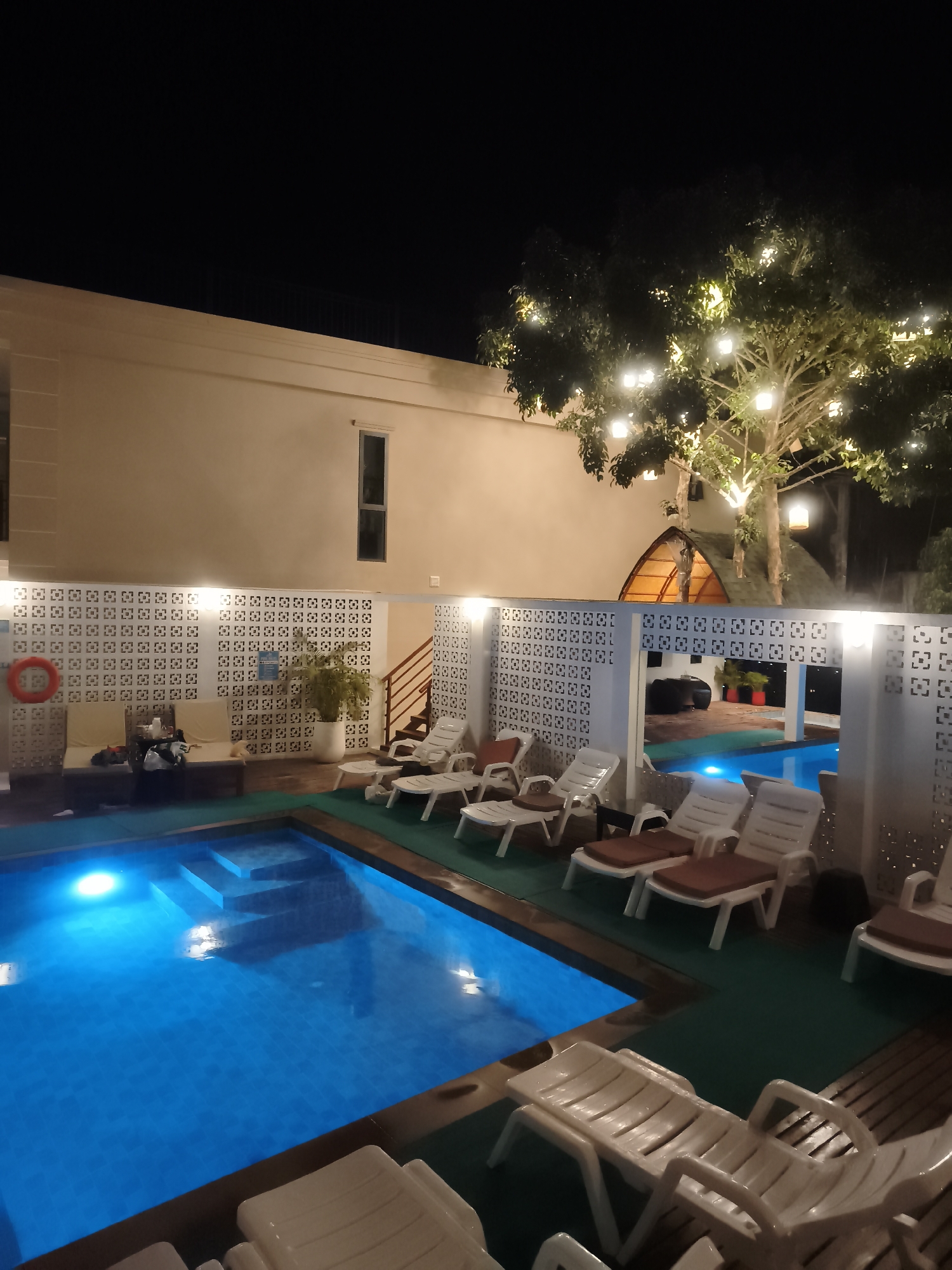 This media file depicts an outdoor swimming pool area at night, likely at a resort or hotel in Ban Rak, Thailand. The scene is illuminated by artificial lights, including lights within the pool that give the water a vibrant blue glow and string lights adorning a tree.
The main subjects are the swimming pool itself, filled with clear blue water and featuring submerged steps, and a collection of white plastic lounge chairs arranged around its perimeter. Some chairs have reddish-brown cushions. In the background, there's a building with a light-colored facade and windows, and a decorative white screen wall with a repeating pattern separates the pool area from the building and other sections of the property. Adjacent to the building, a staircase with wooden steps leads upwards.
A notable detail is the lush green foliage of a tree covered in warm, glowing string lights, adding a festive atmosphere to the setting. The overall mood is tranquil and inviting, suggesting a relaxing evening by the pool. There are no people visible in the frame, and no explicit weather conditions are evident, other than it being dark.
One object visible is a red life preserver ring hanging on a stand. Some bags and what appear to be cushions are placed near the screen wall. A potted plant is visible in a white pot. There is a small, arched structure with a thatched roof partially visible behind the tree, suggesting a unique architectural element of the location. No visible text is discernible in the image, apart from a possible sign on the screen wall which is too blurry to read.