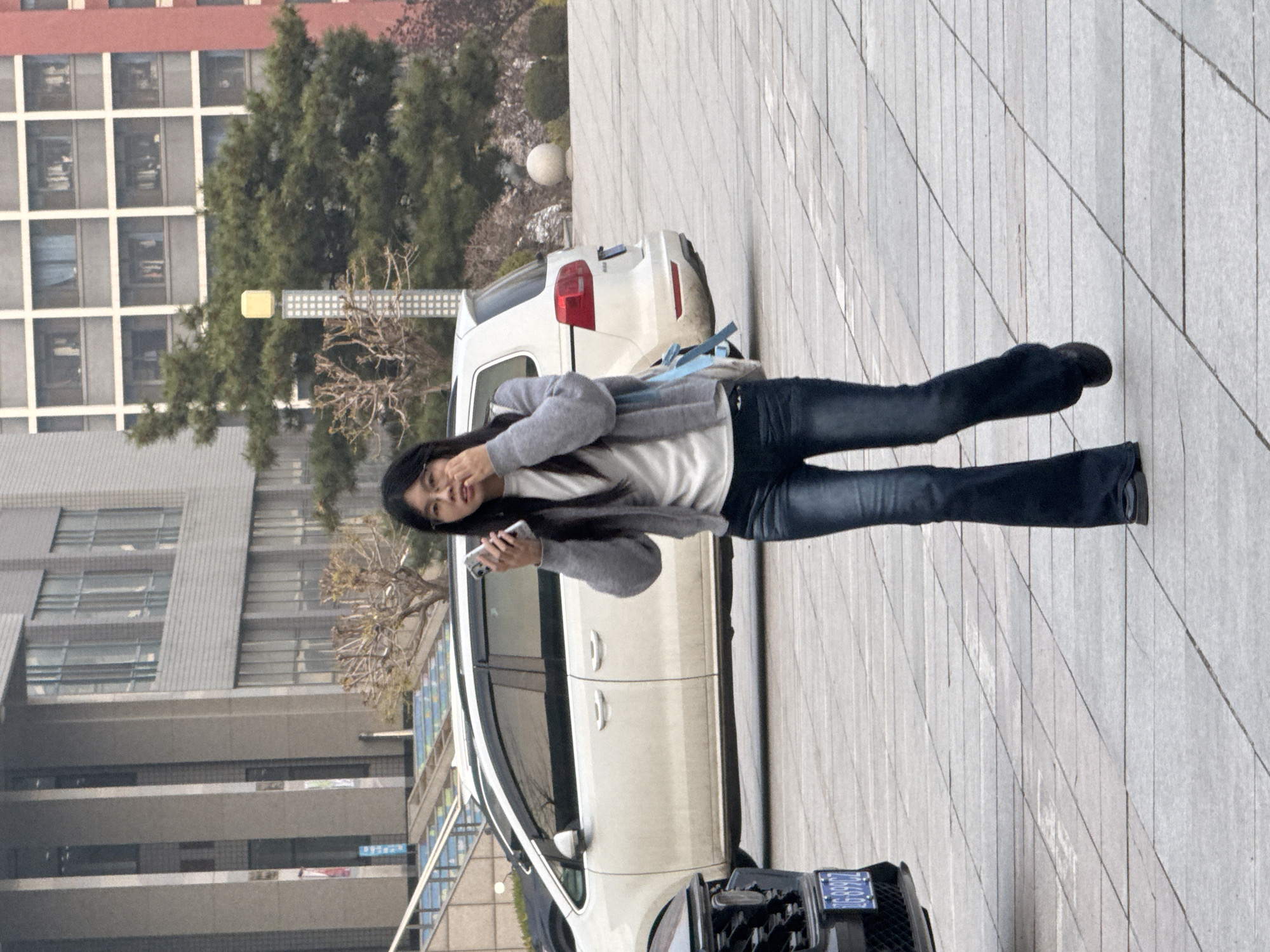 A medium shot shows a woman standing next to a white car, holding a cell phone in her left hand and gesturing with her right. She is wearing glasses, a white shirt, a gray cardigan, and jeans. The white car is parked on a gray, tiled surface. In the background, there are buildings and trees. The license plate on the car is visible, indicating the location is Zhongtun, China.