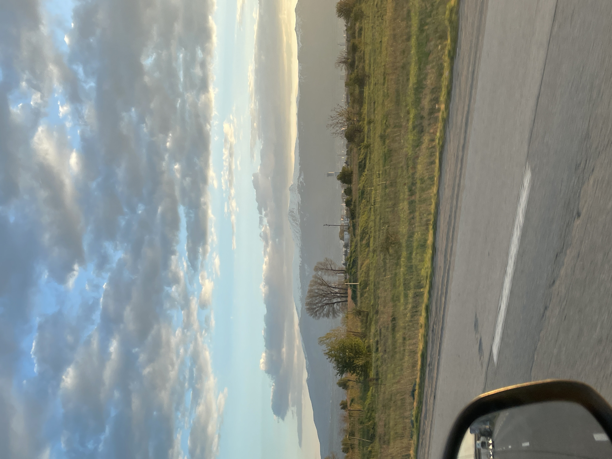 An outdoor, eye-level shot captures a scenic landscape from a moving vehicle, with the image oriented vertically but depicting a horizontal scene. The sky, partially covered by cumulus and stratocumulus clouds, dominates the upper left, displaying a mix of light blue and patches of sunlight illuminating the cloud edges with a soft golden hue. In the mid-ground, a range of mountains stretches across the horizon, with some peaks appearing snow-capped or light-colored due to the sun. The mountains are bathed in a soft, diffused light, suggesting late afternoon or early evening.
To the right of the mountains, a verdant grassy field, possibly a pasture or undeveloped land, extends towards the foreground, separated from the road by what appears to be a fence line. Scattered trees, some bare-branched and others with green foliage, dot the field and the base of the mountains. A white, multi-story structure is faintly visible near the base of the mountains in the distance, alongside other indistinct buildings, indicating some human settlement.
In the foreground, a paved road with a solid white line on its edge is visible, occupying the bottom left quarter of the frame. The smooth texture of the road contrasts with the natural elements. The reflection of the road and potentially the vehicle's interior can be seen in the bottom left corner, where a portion of a car's side mirror is visible, suggesting the photo was taken from inside a car. The overall impression is one of travel through a peaceful, semi-rural, or mountainous region under a dynamic sky at sunset or sunrise. No visible text is present.