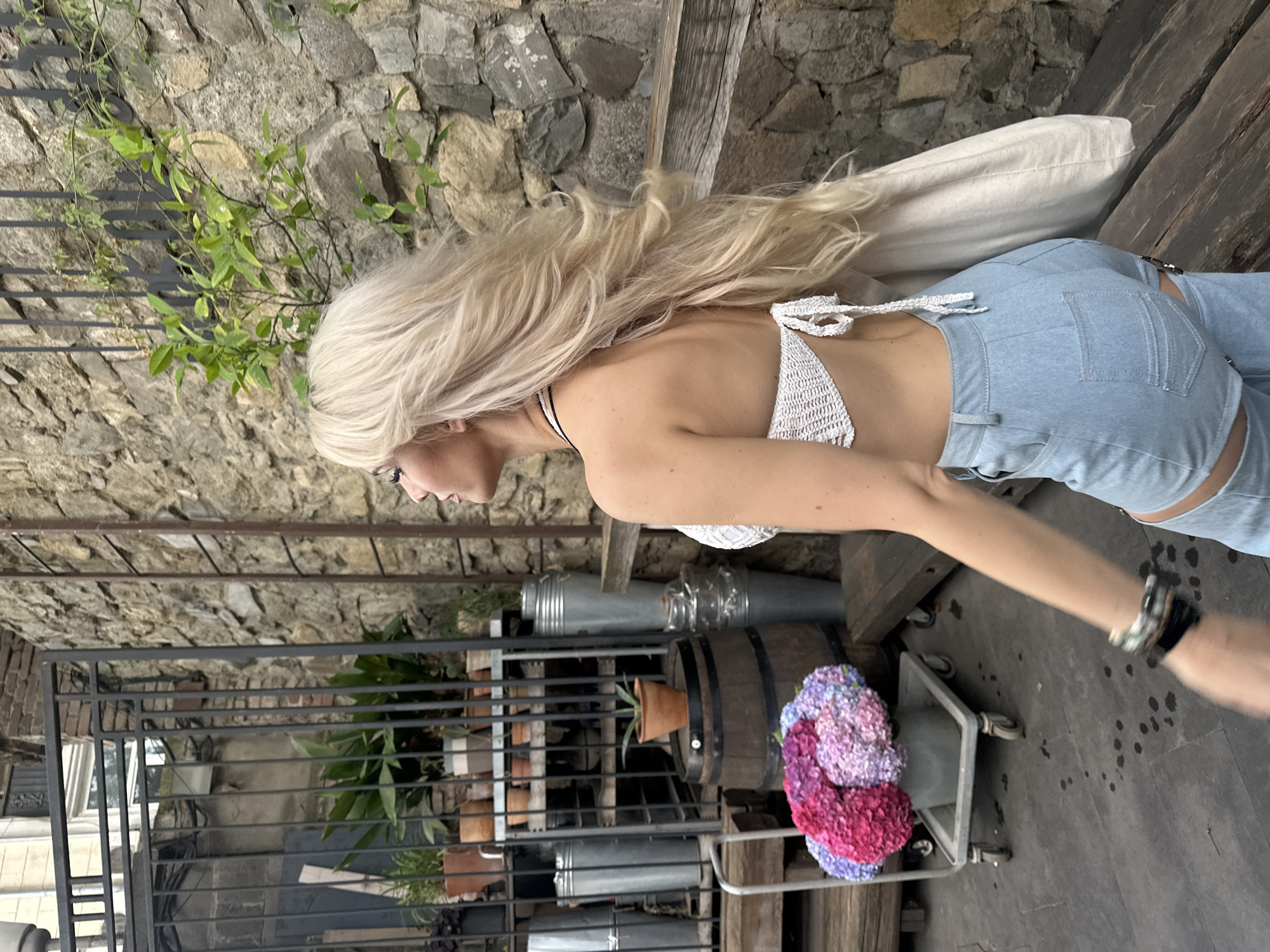A young woman with long, blonde hair, wearing a white crochet top and light blue denim shorts, walks past a stone wall and various plants in what appears to be an outdoor courtyard or garden area in Tbilisi, Georgia. The woman is captured in profile, looking to her left, with her hair blowing in the wind. In the background, there is a metal fence, shelves with potted plants, a wooden barrel, and large galvanized metal containers. To the left of the woman, a cart holds a large bouquet of pink and purple hydrangeas. The ground is paved with dark stones, showing some wet spots. The overall scene suggests a warm, possibly summer day.