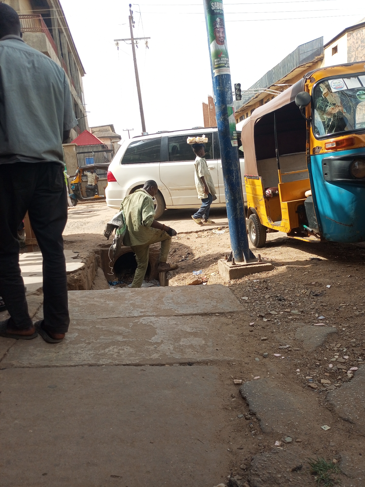 The image shows a street scene in Maiburiji, Nigeria. A man dressed in a green uniform is knee-deep in an open manhole, seemingly working on the infrastructure. To his left, another man is standing on the sidewalk, observing the scene. In the background, a white van and a yellow and blue auto-rickshaw (tuk-tuk) are parked along the street. A young boy carries a tray on his head as he walks past the parked van. A utility pole with a political poster is visible near the auto-rickshaw. The street surface is a mix of concrete and dirt, showing signs of wear and tear. Buildings are visible in the background, suggesting an urban setting.