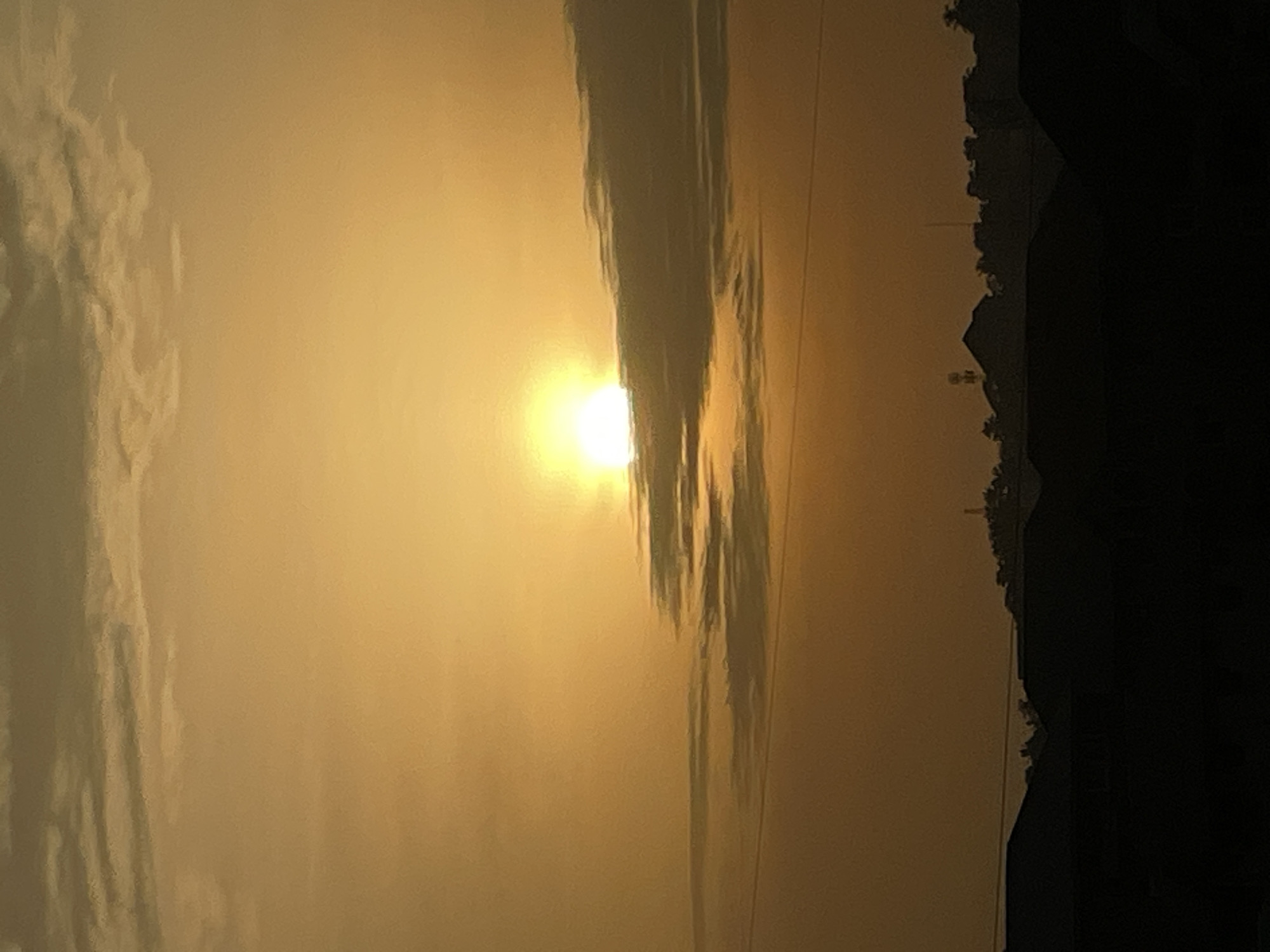 The image shows a horizontal view of Nkpor, Nigeria at sunrise/sunset. The sun is partially obscured by a dark cloud formation. To the right, tree silhouettes and building tops are visible against the bright sky. The overall light is a hazy, golden hue.
