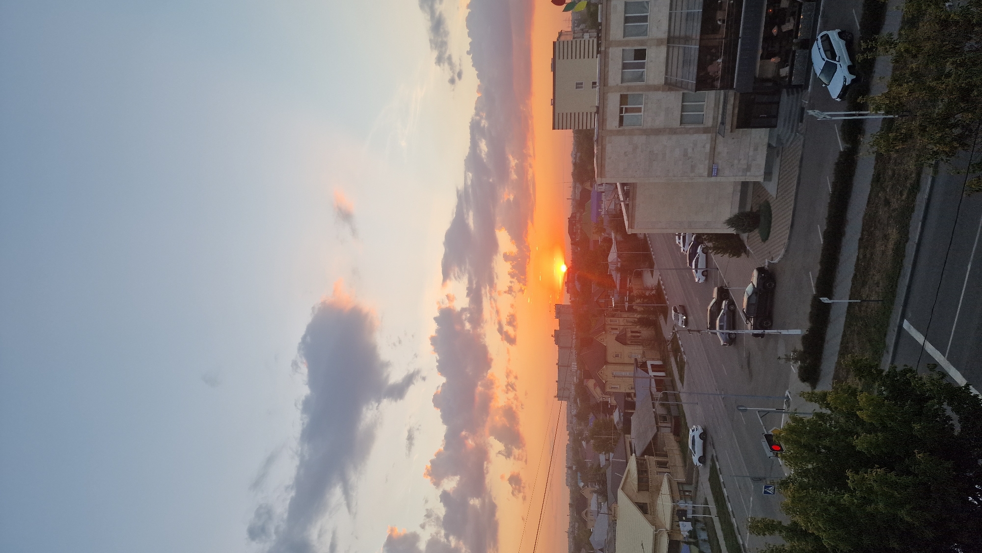 This is a photograph taken from an elevated vantage point in the city of Kostanay, Kazakhstan, during sunset. The sky is a gradient of pale blue to a warm orange, illuminated by the setting sun which is positioned near the horizon, partially obscured by buildings and clouds. Scattered clouds are visible, some tinged with orange and pink from the sunset.
In the foreground, a street intersection is visible. Several cars are present on the road, including a black pickup truck, a white SUV, and other sedans. A traffic light shows a red signal. On the left side of the intersection, a prominent green tree is in the foreground. Across the street, there are several buildings, including a multi-story office or apartment building with a modern facade and awnings over outdoor seating areas. Smaller residential buildings with distinct rooflines are also visible in the mid-ground. The scene appears to be a typical urban or suburban environment in Kostanay. The time of day is clearly late afternoon or early evening, as indicated by the low angle of the sun and the warm lighting.
