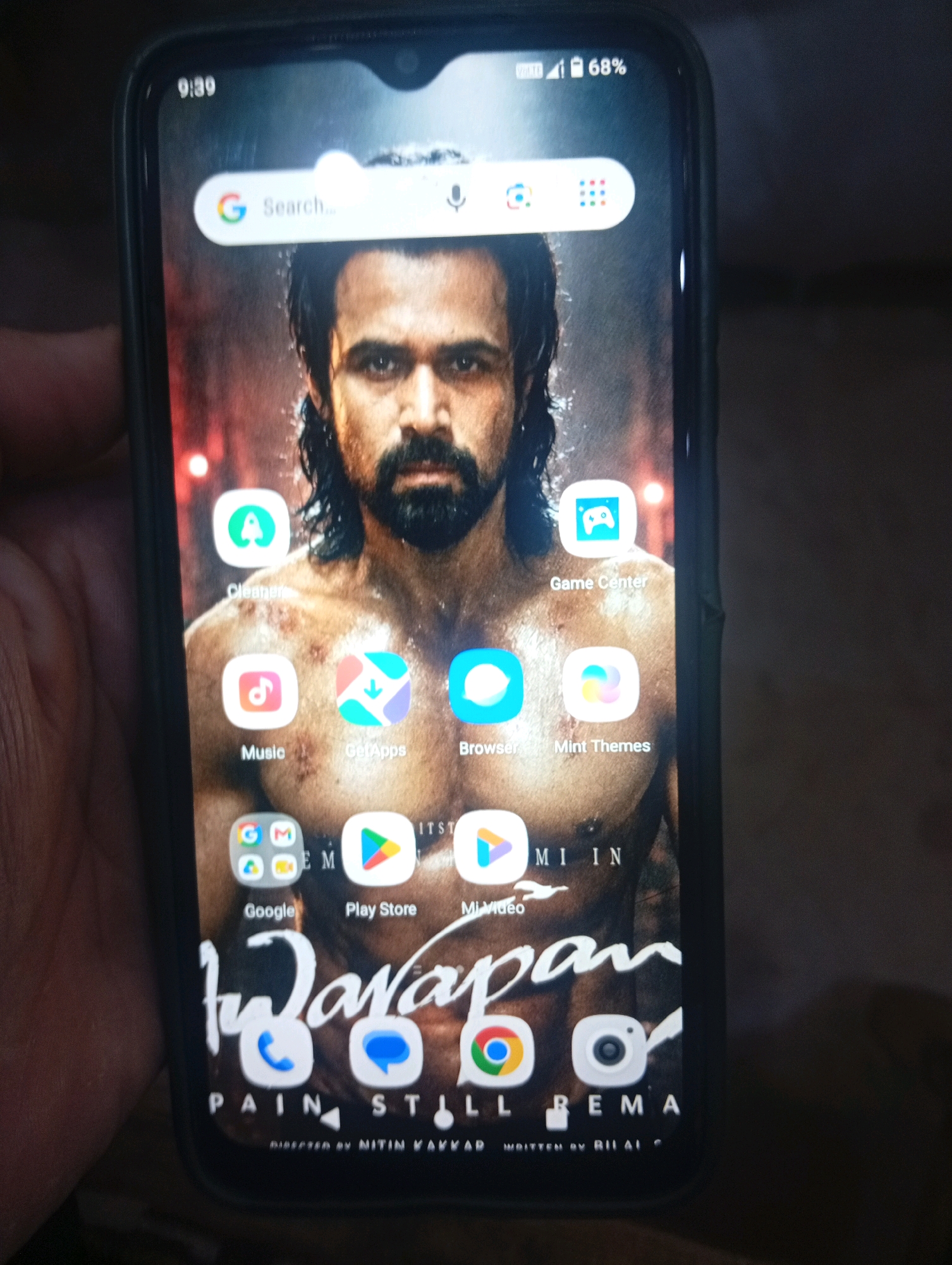 The image shows a smartphone with its screen on, displaying a wallpaper and app icons. The wallpaper features a close-up of a man's face and upper body, with a dark, fiery background. Prominently displayed on the screen is a search bar labeled "Search..." with a Google "G" logo. Below the search bar are rows of app icons, including "Cleaner," "Game Center," "Music," "GetApps," "Browser," "Mint Themes," "Google," "Play Store," and "MiVideo." At the bottom of the screen, the title of what appears to be a movie or show, "Warapan," is visible, along with text that seems to be credits. The phone is being held by a hand on the left side, partially obscuring the screen. The ambient lighting suggests the photo was taken indoors, possibly in a dimly lit room. There is no indication of the city of Larkana, Pakistan, in the image.