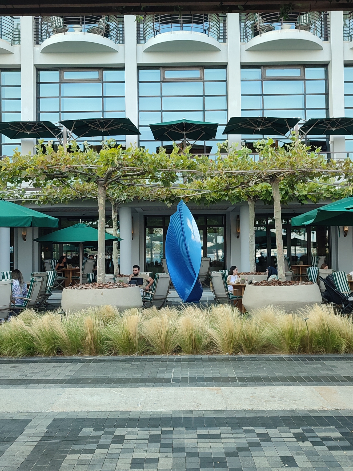 The image captures an outdoor seating area of a restaurant or cafe, likely located in Istanbul, Turkey, given the architectural style and presence of elements common to waterfront establishments. In the foreground, a paved walkway with a geometric pattern of dark and light grey tiles is visible. Beyond the walkway, a dense arrangement of tall, feathery ornamental grass in hues of beige and green creates a naturalistic border.
Behind the grass, a row of outdoor tables and chairs are set up under large, dark green umbrellas, suggesting a shaded dining area. Several trees with sparse foliage are planted in large, light-colored concrete planters, providing additional shade and contributing to the ambiance. A striking blue, abstract sculpture with a flowing, ribbed design stands prominently on the left side of the scene, adding a contemporary artistic touch.
Several patrons are seated at the tables, engaged in conversation or appearing relaxed. On the far left, a woman in a patterned dress is seated. Further into the scene, a man is seen sitting at a table with a laptop. Another woman, wearing a hijab, is seated at a table with other individuals. In the background, the facade of a modern building with multiple floors is visible. The building features large, grid-like windows reflecting the sky and a series of balconies with curved railings on the upper levels. The overall scene suggests a pleasant outdoor dining experience in a modern urban setting.