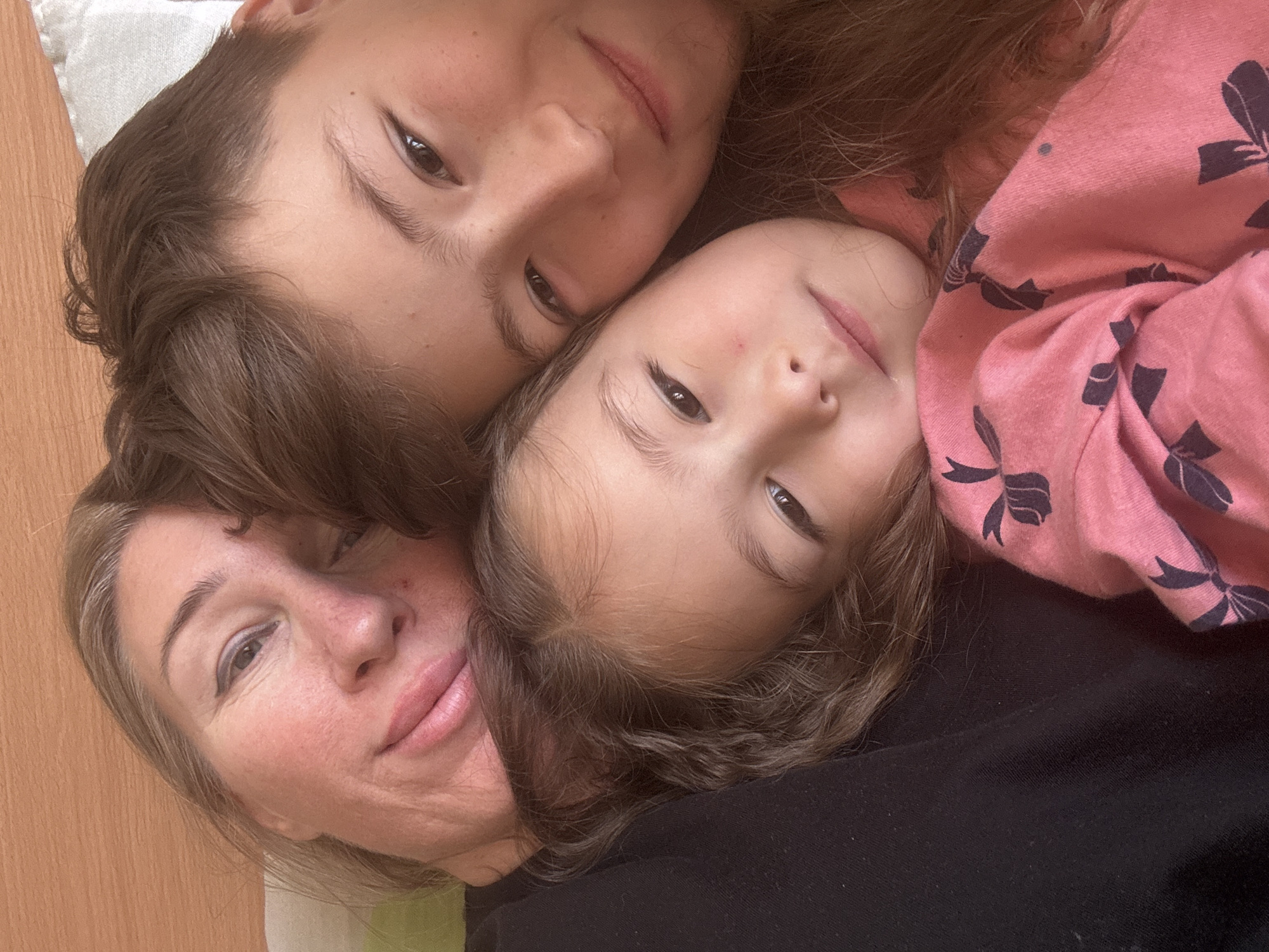 A selfie taken indoors shows an adult woman and two young girls in close proximity. The woman has fair skin, light brown hair with streaks of gray, and is wearing makeup. One girl is wearing a pink top with a bow pattern. The second girl has brown hair and is positioned between the woman and the first girl. The background appears to be a plain wooden surface. The location is not evident from the image. The city of Tashkent, Uzbekistan cannot be confirmed.