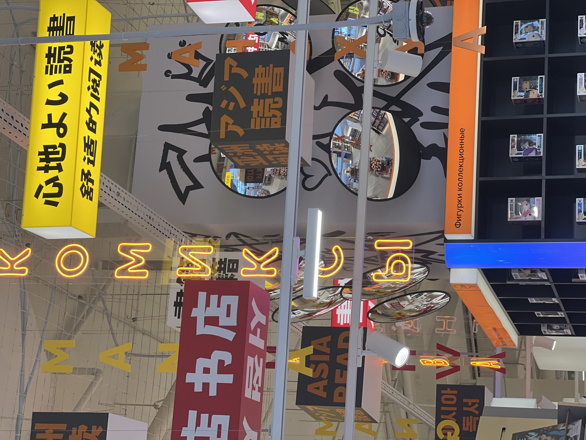 The image shows the interior of a retail store, likely a bookstore or a store selling merchandise. Various signs and displays are suspended from the ceiling. In the upper part of the image, a yellow sign with Japanese and Chinese characters is visible, reading "心地よい読書 舒适的阅读" which translates to "Comfortable Reading". Below this, several large, yellow neon letters form the word "KOMM" and "K". A red sign with white Chinese characters hangs prominently, reading "书店", meaning "bookstore". Other signs with yellow and black lettering, including "ASIA READ", are also present. Multiple circular mirrors are hung at different levels, reflecting parts of the store's interior, showing shelves stocked with various items and what appear to be more signs. The ceiling is a grid of white beams and suspended lighting fixtures. In the lower part of the image, a black shelving unit is filled with collectible figures in boxes, and an orange sign above it reads "Фигурки коллекционные" in Russian, meaning "Collectible figures". The overall impression is a vibrant and organized retail environment. There are no visible people in the image. The style of the signage and the presence of collectible figures suggest a modern retail space that might be found in a city like Saint Petersburg, Russia, given the Russian text.