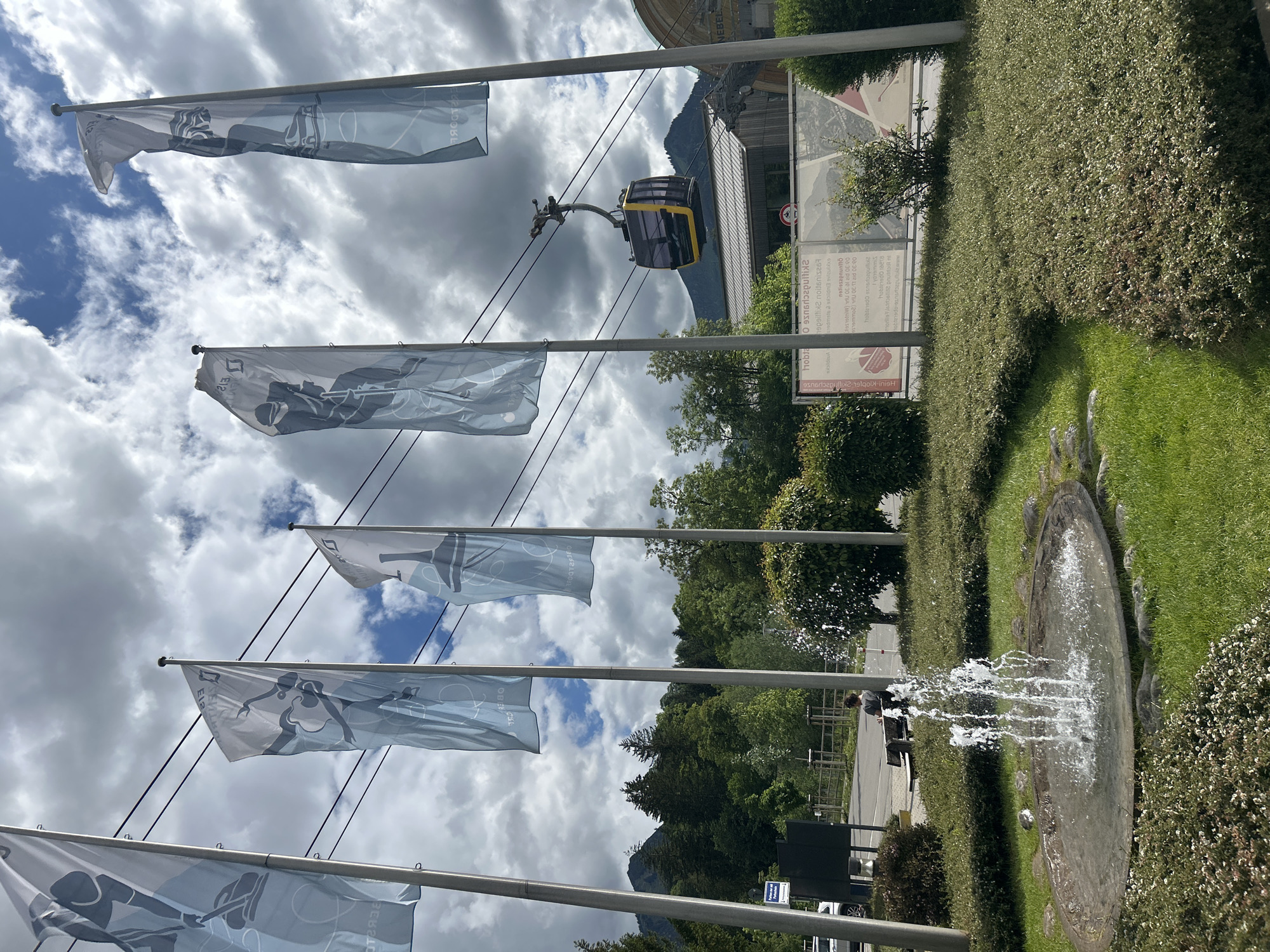 The image depicts the entrance area of a cable car station in Oberstdorf, Germany, likely the Nebelhorn cable car. In the foreground, a circular ground-level fountain with multiple water jets is situated within a manicured grassy area, interspersed with small rocks and low-lying green bushes.
Five vertical flags on tall, light grey poles dominate the midground. These flags feature light blue and white designs depicting stylized figures engaged in various winter sports such as curling, ice hockey, and bobsledding or skeleton. The text "OBERSTDORF" and "EISSPORT" is visible on several flags, often mirrored, suggesting the image is captured from a perspective where the flags' reverse side is visible.
Behind the flags and bushes, a paved area, potentially a road or parking lot, is visible. A person in a dark shirt and pants is seated on a wooden bench to the left of the fountain, facing away from the camera. Further back on the right, a large billboard-like sign displays mirrored German text, including "Skiticket" (ski ticket) and "Öffnungszeiten" (opening hours), along with numerical operating times such as "09:00 - 16:00," indicating an information point for a ski or mountain facility.
An yellow and black cable car gondola is suspended on cables overhead, traversing towards a modern building with a wooden facade and large windows, identified as the cable car station, situated in the upper midground. Dense green trees, a mix of coniferous and deciduous, cover the slopes of mountains that rise in the background, particularly on the left and right sides of the frame. The sky above is partially overcast with white and grey clouds, interspersed with patches of blue sky.