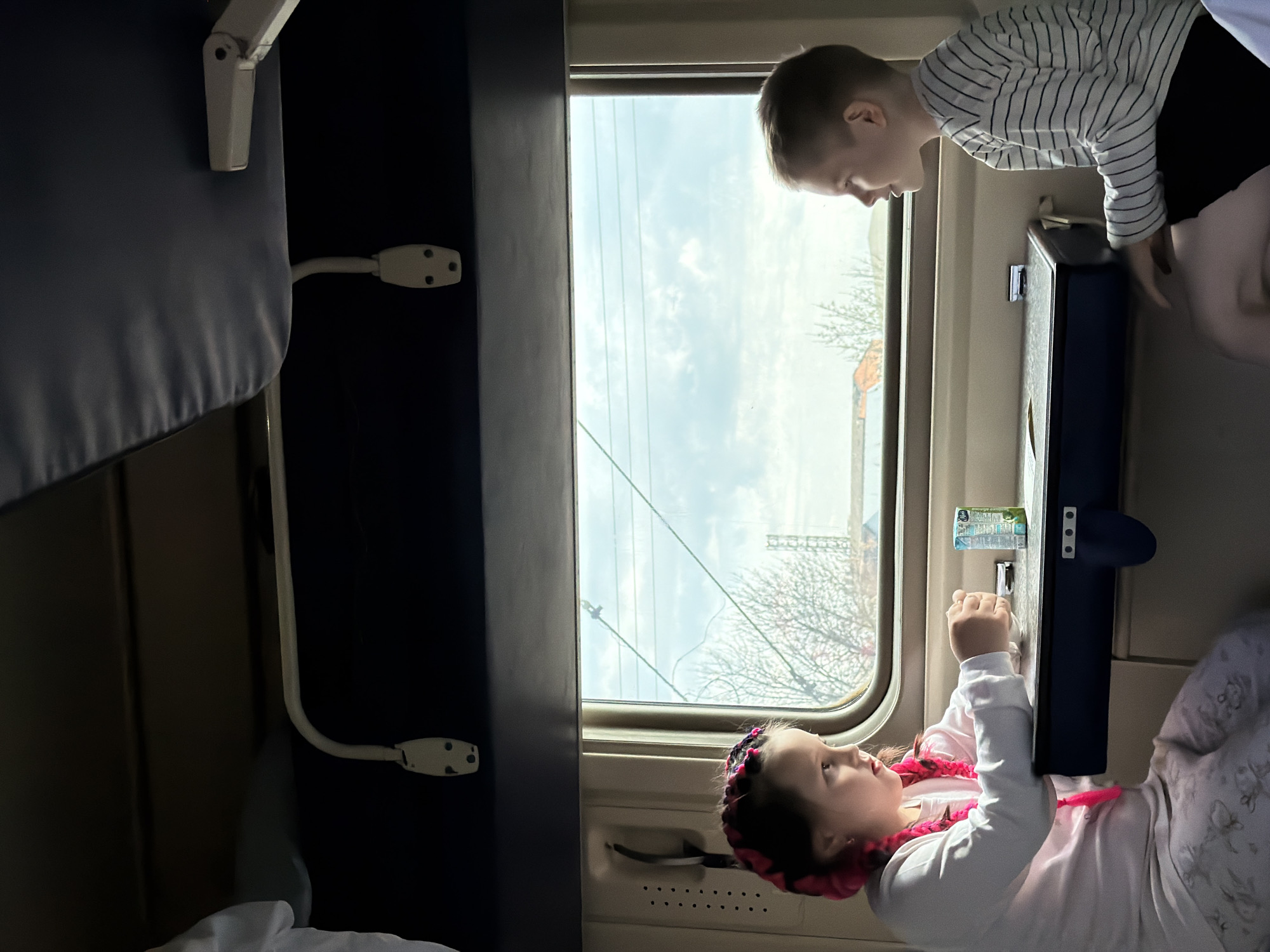 Two children, a boy and a girl, are in a train compartment. The boy is standing and leaning over the small table attached to the wall near the window. The girl is lying down, looking at something on the table, possibly a small package. Outside the window, bare trees are visible, suggesting it may be winter or early spring. The scene is likely in the city of Medvyodovskaya, Russia. The interior of the train compartment includes a bed on the left side of the frame.