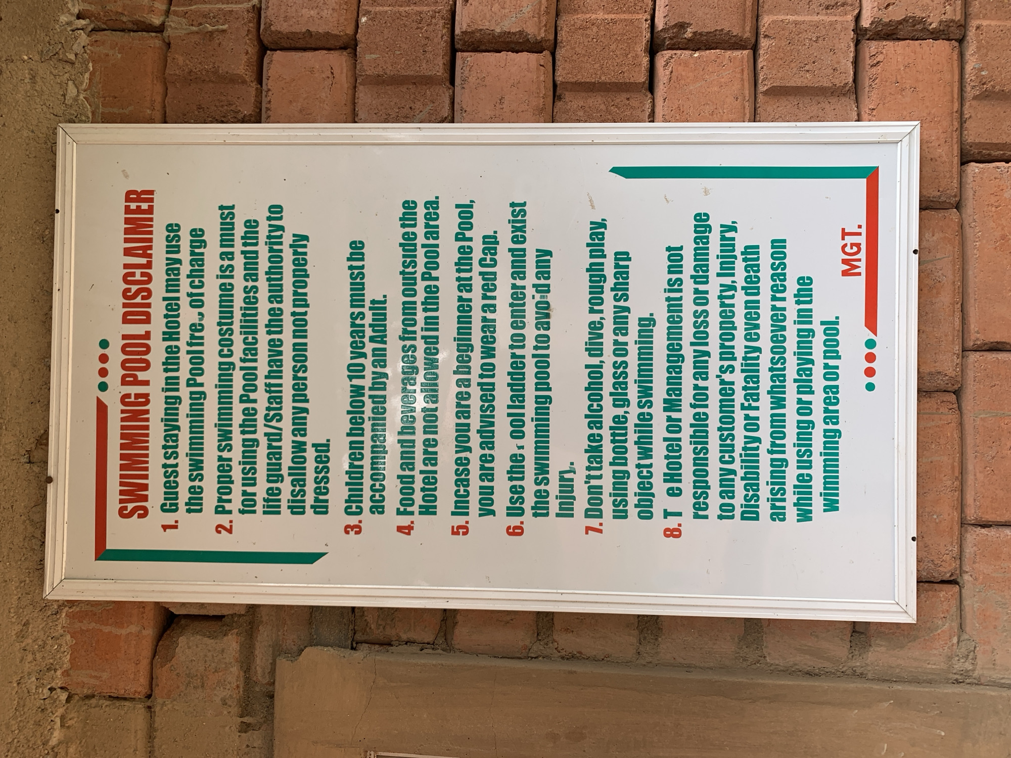 The image shows a white sign with red and green lettering posted on a brick wall. The sign is a "SWIMMING POOL DISCLAIMER" and lists 8 rules for guests using the pool. The sign is framed by a white border. The brick wall is made of reddish-brown bricks. The sign is the main subject of the image. The environment appears to be outdoors, possibly at a hotel or resort. The sign is in good condition, with no visible damage. The lettering is clear and easy to read. The location is not provided in the image.