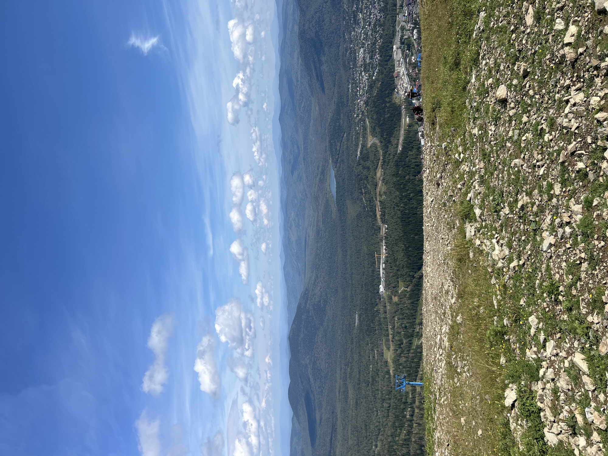 This is a high-angle, wide-angle shot of a mountainous landscape in Sheregesh, Russia. The foreground is a rocky, grassy slope dotted with small yellow flowers. A blue ski lift tower is visible on the left side of the frame. In the mid-ground, a dense, dark green forest covers rolling hills, with a small lake visible in the distance. A town with buildings and infrastructure, possibly a ski resort area, is nestled in the valley. In the far distance, multiple layers of hazy blue mountains stretch across the horizon. The sky is a bright blue with scattered white cumulus clouds. Several people are visible on the slope in the middle ground, appearing to be hiking or walking. The scene appears to be set on a clear, sunny day.