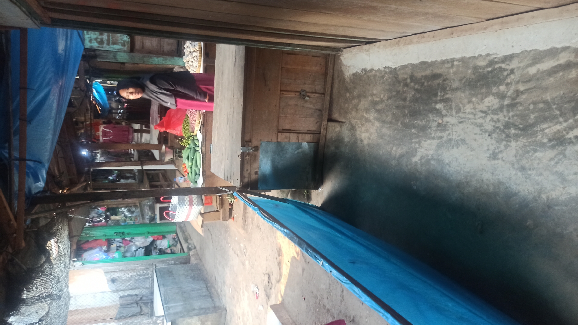 A woman wearing a black hijab and a pink dress stands behind a market stall laden with produce in Trisinar, Indonesia. The stall is covered by a blue tarp, and wooden planks form the stall's structure and flooring. In front of the stall, a blue tarp hangs diagonally, obscuring the ground. The background reveals more market stalls and some clothing racks. The ground is dirt and concrete.