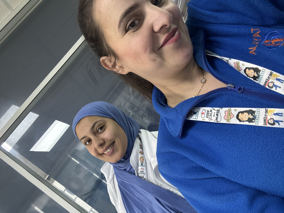 The image displays two adult women posing for a selfie indoors. The woman in the foreground on the right has dark hair pulled back and is smiling directly at the camera. She is wearing a blue fleece zip-up jacket. A white lanyard with various cartoon figures and text such as "KEEP CALM I'M A NURSE," "MCSTEAMY," "CALLIE," and "DANCE IT OUT" is visible around her neck. A gold chain necklace with a white four-leaf clover pendant is also worn. The name "ALIYAT" and a logo resembling a stylized "B" or "D" with a cross are embroidered on the left chest area of her jacket. The woman in the background on the left is also smiling at the camera and wears a light blue hijab over a white garment, possibly a uniform top, with a lanyard visible underneath. The background shows a reflective surface, likely a window or glass partition, revealing reflections of interior lighting and horizontal blinds. The specific location within the city of Meknes, Morocco, cannot be definitively determined from the visual information provided; however, the attire and lanyard text suggest a professional setting such as a medical facility or educational institution.