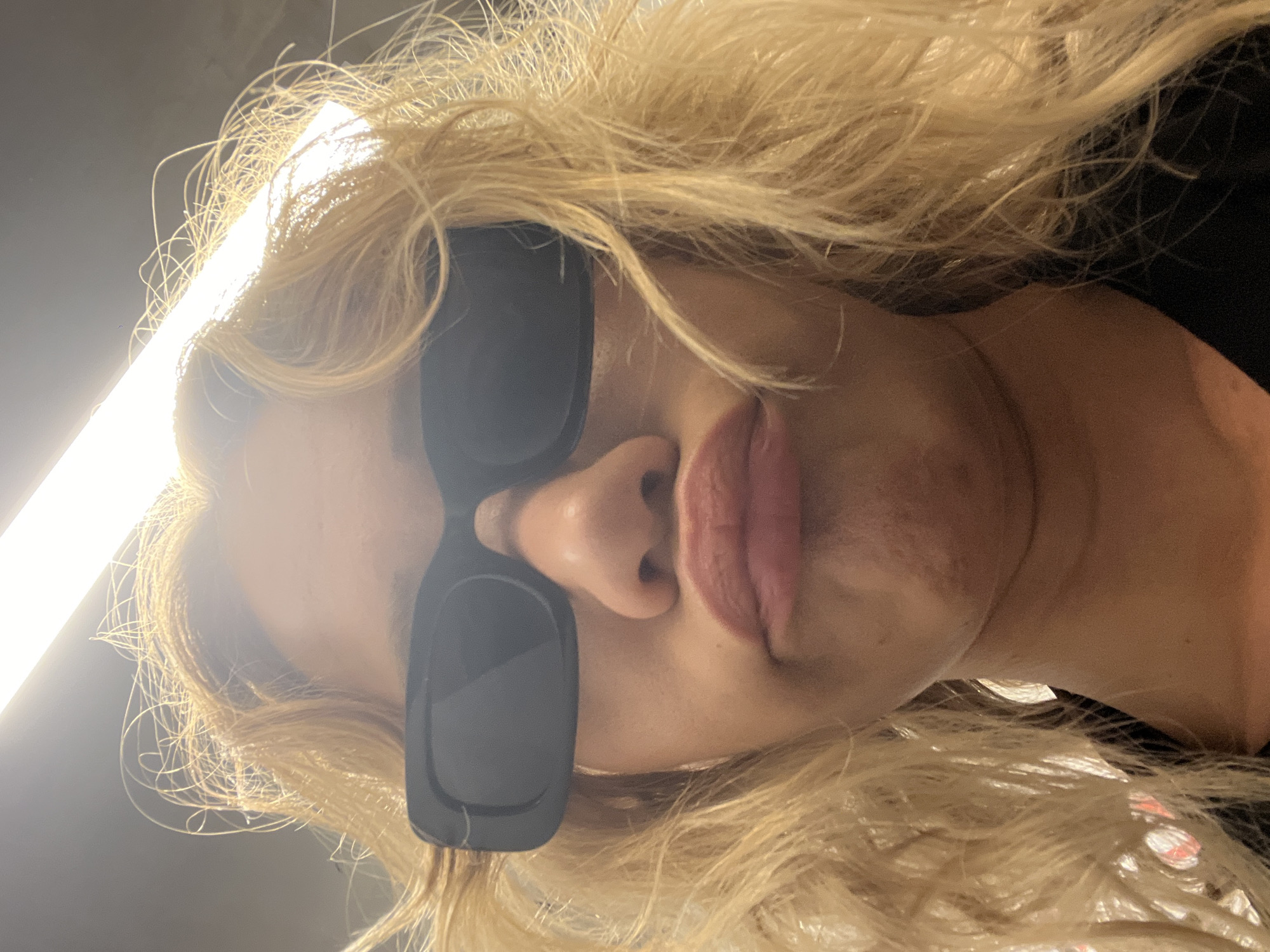The media file is a close-up, low-angle shot of a person's face, with their head tilted downwards. The person has blonde hair, which is styled in loose waves and appears to be illuminated by a bright light source from the left. They are wearing dark, rectangular sunglasses that cover a significant portion of their face. Only their nose and lips are clearly visible below the sunglasses. The lips are full and appear to be painted with a pinkish-red lipstick. The skin tone is light, and there are some subtle shadows and highlights visible on the face, suggesting indoor lighting. The background is dark and out of focus, with a bright, linear light source on the left side of the frame. There is no discernible text in the image. The overall impression is of a casual, perhaps candid, shot taken from an unusual perspective. The location context of Mühldorf a. Inn, Germany, is not visually evident from the image itself.