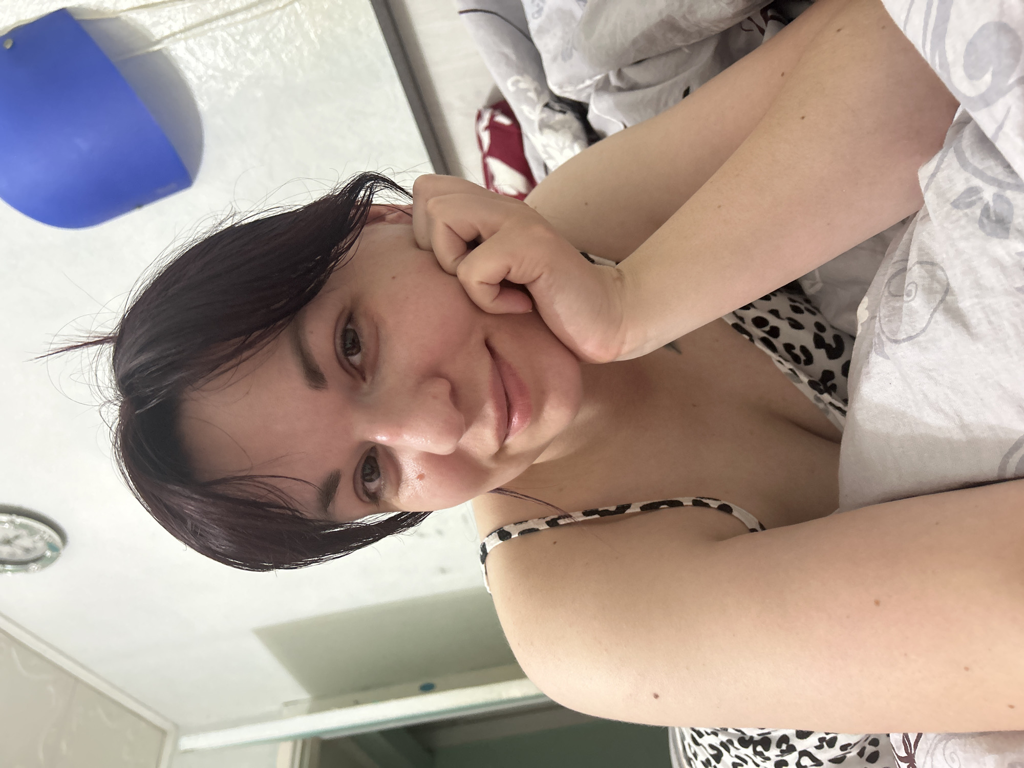 A young woman is lying in bed, propped up on her left elbow with her right hand supporting her chin. She is looking directly at the camera with a slight smile. Her dark hair is pulled back, with a few wisps falling forward around her face. She is wearing a black and white leopard print camisole. The bed has white and gray patterned sheets and a red and white patterned pillow is visible behind her. The room appears to be a bedroom, with a white wall behind the bed. A clock is visible on the wall in the upper left corner. A blue object, possibly a lamp or light fixture, is attached to the wall on the right side of the image. The overall impression is one of a casual, personal moment captured in a bedroom. There is no indication of the city of Фоки, Russia in the image itself.