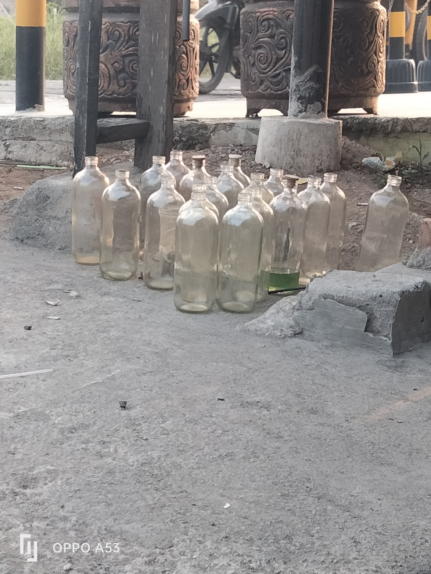 The image depicts a collection of empty glass bottles and one bottle containing a green liquid, arranged on a concrete surface. The bottles appear to be arranged in a scattered manner, with some standing upright and others leaning. In the background, there are large decorative pots, wooden posts, and parts of a motorcycle. The scene suggests an outdoor environment, possibly a roadside or a temporary storage area. The presence of the decorative pots and the general setting could indicate a location in Karawang, Indonesia, known for its cultural heritage and industrial zones.