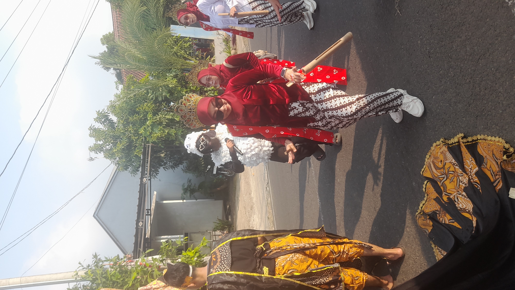 This is a medium shot, eye-level outdoor photograph taken in Lemahabang, Indonesia, likely during a festival or parade. The scene is set on a paved road with houses and trees in the background. The sun is casting long shadows on the road, indicating it is late afternoon.
In the foreground, a person dressed in traditional Indonesian attire, including a red velvet top, a red and white patterned sarong, and a red hijab, is holding a bamboo prop. The person is wearing sunglasses and has their hands in a gesturing pose. Next to them, another person in similar traditional red attire is partially visible.
Further back, a person is in a sheep costume with a fluffy white body and black face, holding a microphone. Behind them, another individual is seen from the back, wearing a black robe with yellow trim and patterned orange pants. The costume and attire suggest a cultural performance.
The environment includes a street, houses with pitched roofs, and lush green trees. The overall atmosphere is festive and celebratory.