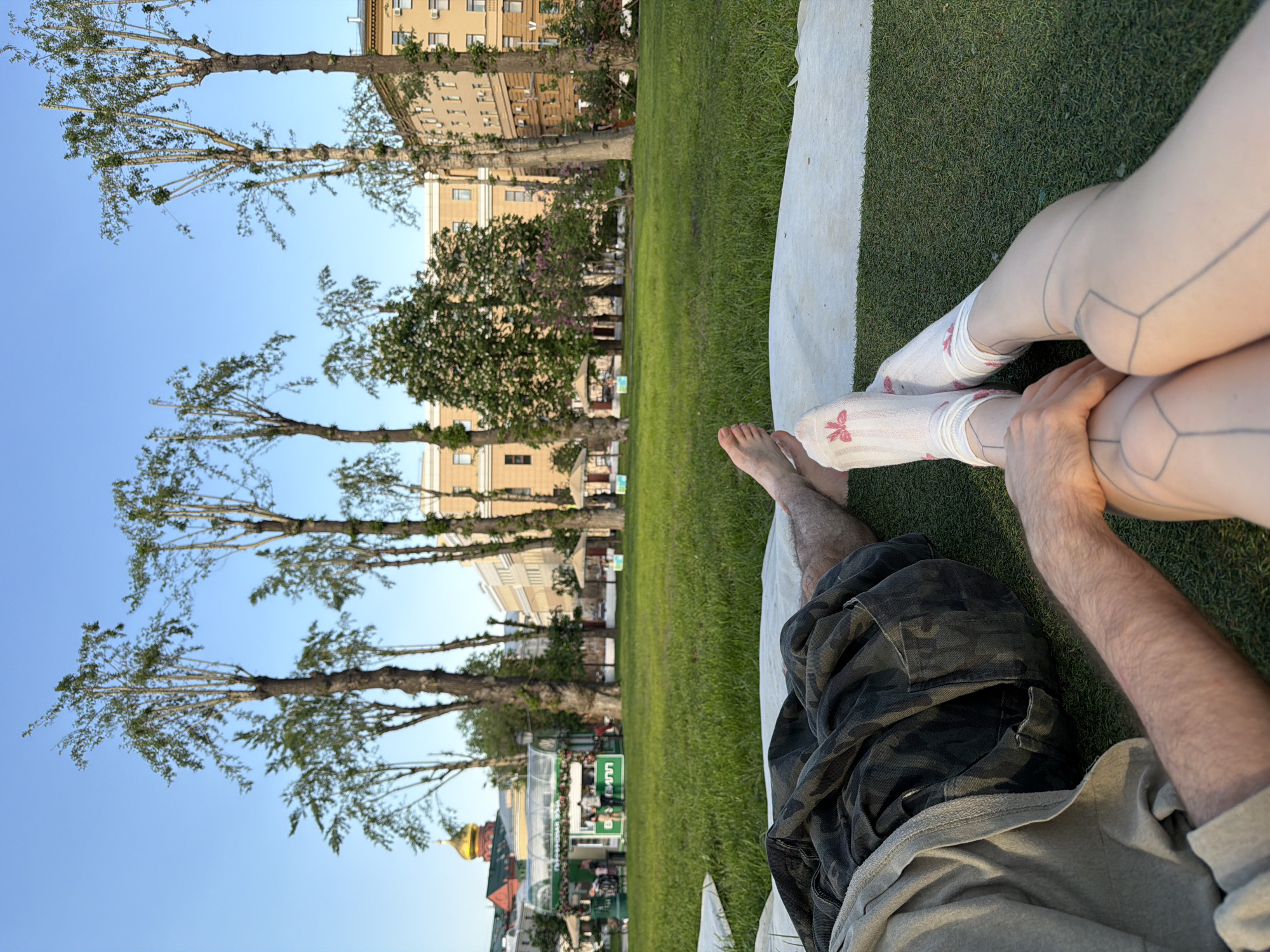 The image shows a view of a couple lying on a blanket in Zaryadye Park in Moscow, Russia. The man is wearing camouflage shorts and a gray shirt, while the woman is wearing patterned stockings. They are holding hands. The view behind them shows the park's green grass, trees, and building architecture, with a glimpse of the Moscow skyline.
