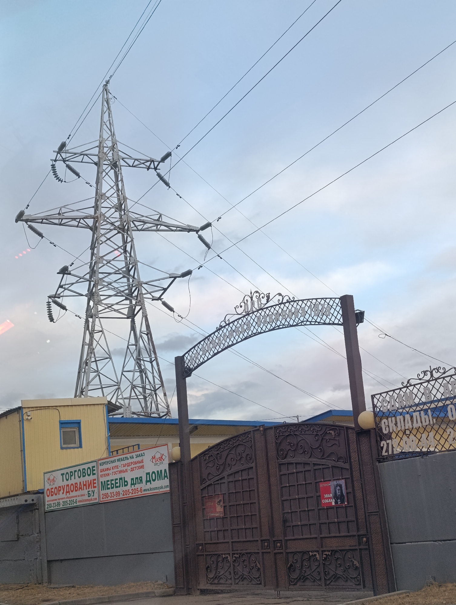 The image depicts an industrial or commercial area in Krasnoyarsk, Russia, under an overcast sky during daytime. A large metal electricity pylon dominates the left side of the frame, with numerous power lines extending across the sky, some attached to the pylon via insulators.
In the foreground, an ornate, dark brown metal gate with a decorative archway opens into a property. To the left of the gate is a utilitarian yellow building with a blue trim, possibly a modular office or storage unit. Attached to this building is a large white sign with text in Russian.
Visible text on the sign indicates a business related to "ТОРГОВОЕ ОБОРУДОВАНИЕ" (Commercial Equipment), including "Оборудование на заказ" (Equipment by order) and "Б/У" (Used). It also advertises "МЕБЕЛЬ ДЛЯ ДОМА" (Furniture for Home) and "КОРПУСНАЯ МЕБЕЛЬ НА ЗАКАЗ" (Cabinet Furniture by Order), specifically "ШКАФЫ КУПЕ • ГАРДЕРОБНЫЕ" (Wardrobes • Dressing rooms), "КУХНИ • ГОСТИНЫЕ • ДЕТСКИЕ" (Kitchens • Living rooms • Children's rooms). The company name "МАЯК" (Mayak) and website "www.krasmayak.com" are also visible, along with a phone number (250-33-99-205-205-6).
On the right leaf of the gate, there's a red and white sign with the text "ОСТОРОЖНО ЗЛАЯ СОБАКА" (Beware, Evil Dog), featuring an image of a dog. To the far right, another white sign on a fence partially reads "СКЛАДЫ, О" (Warehouses, O) and "27". The decorative text on the archway above the gate is hard to decipher but appears to be a business name. The overall impression is an active commercial or storage facility.