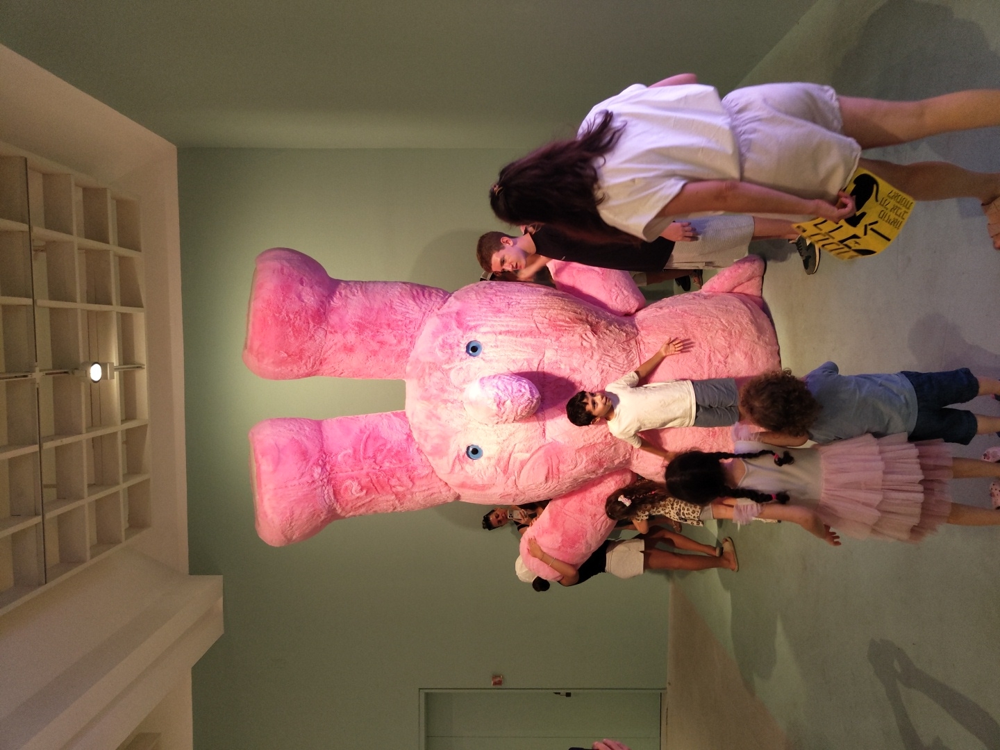 The image depicts a large, pink, plush bunny sculpture in an indoor exhibition space. Several people, including adults and children, are interacting with or observing the sculpture. The environment appears to be an art gallery or museum, characterized by its neutral-colored walls and ceiling with a grid-like structure. The lighting is artificial and evenly distributed.
Specifically, the scene includes:
* **The Sculpture:** A very large, fuzzy, pink bunny with prominent ears and blue eyes. It is the central focus of the image.
* **People:**
* A young boy in a white t-shirt and grey shorts stands near the sculpture's lower body, appearing to be interacting with it.
* Two young girls in the foreground, one with pigtails and wearing a pink dress, the other with curly hair in a blue shirt and dark shorts, are looking at the sculpture.
* An adult woman in a white outfit is in the right foreground, holding a yellow sign with text.
* Other adults are visible around the sculpture, some partially obscured.
* **Environment:**
* The walls are painted a pale green.
* The ceiling is white with a visible grid structure and recessed lighting.
* The floor is a smooth, light-colored surface.
* A door is visible on the left side of the frame.
The yellow sign held by the woman in white has Hebrew text on it, suggesting the location may be in Israel. While the image itself does not definitively place it in Tel-Aviv, an art exhibition featuring a large, whimsical sculpture like this is consistent with venues found in major cities like Tel-Aviv.