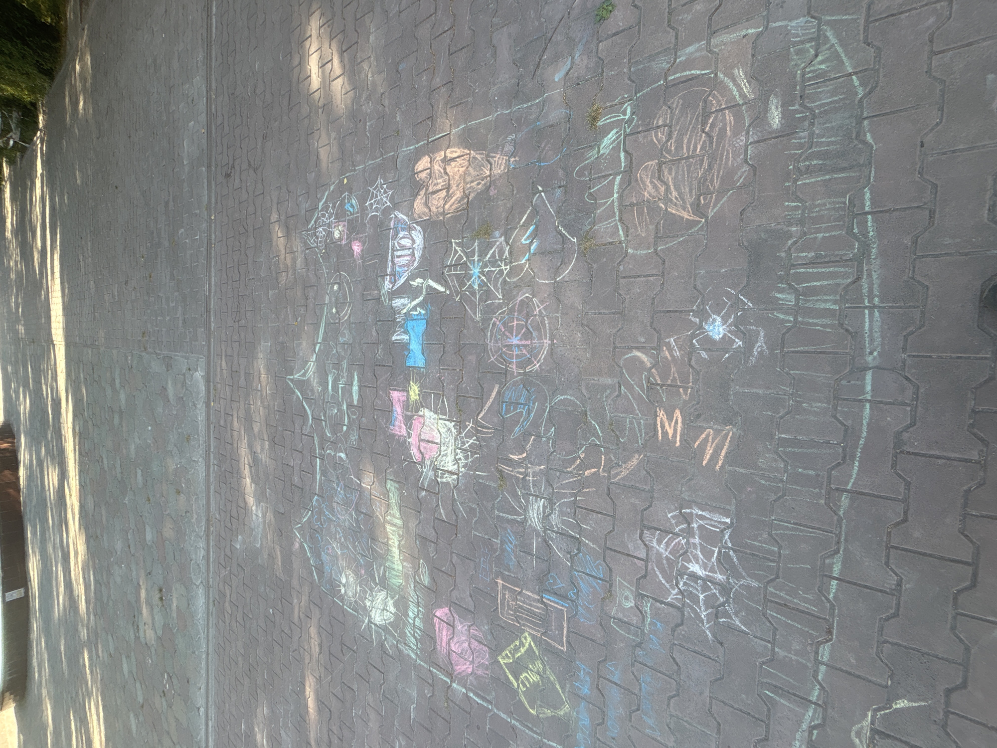 The image shows a chalk drawing on a paved surface in the city of Ob, Russia. The drawing is colorful and appears to be a collaborative effort, with various shapes and figures, including what looks like a house, a heart, spiderwebs, and abstract patterns. The chalk colors used are pink, blue, green, yellow, and orange. The paving stones are interlocking and appear to be made of concrete. Sunlight casts shadows across the drawing and the ground, indicating it is daytime. The background shows more paving stones and hints of greenery and trees. The scene is outdoors, likely in a public space such as a park or sidewalk.