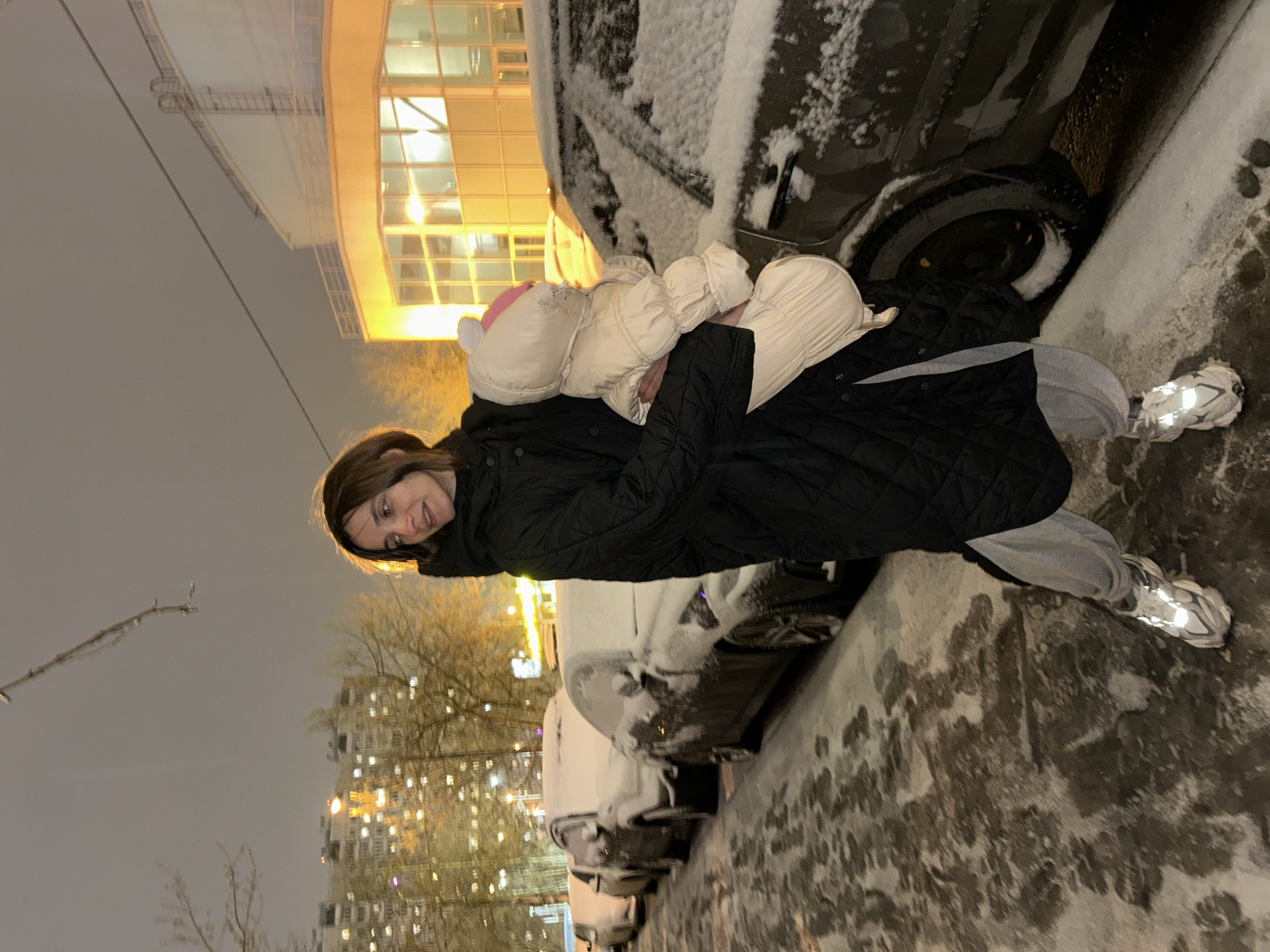 A woman holds a child in front of snow-covered cars in Moscow, Russia. The woman wears a black quilted jacket, gray pants, and white sneakers. The child is dressed in a light-colored snowsuit and pink hat with bunny ears. The cars are heavily covered with snow. In the background, apartment buildings and a brightly lit building with curved architecture can be seen. The environment suggests it is nighttime with snow on the ground.