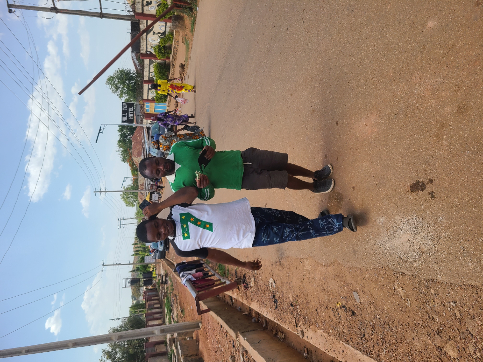 Two men stand on a paved street in the city of Unknown, Unknown. The man on the left wears a white t-shirt with a large green numeral "7" and several yellow stars on it, along with dark pants. The man on the right wears a green shirt and dark shorts. Both men have dark hair and skin. In the background, there is a sign for "HILL TOP HOTEL" and several people are walking down the street. Overhead power lines run above the street. To the left of the men, there is a concrete drainage ditch and some miscellaneous items, including what appears to be a ladder with clothes on it. The sky is blue with some clouds.