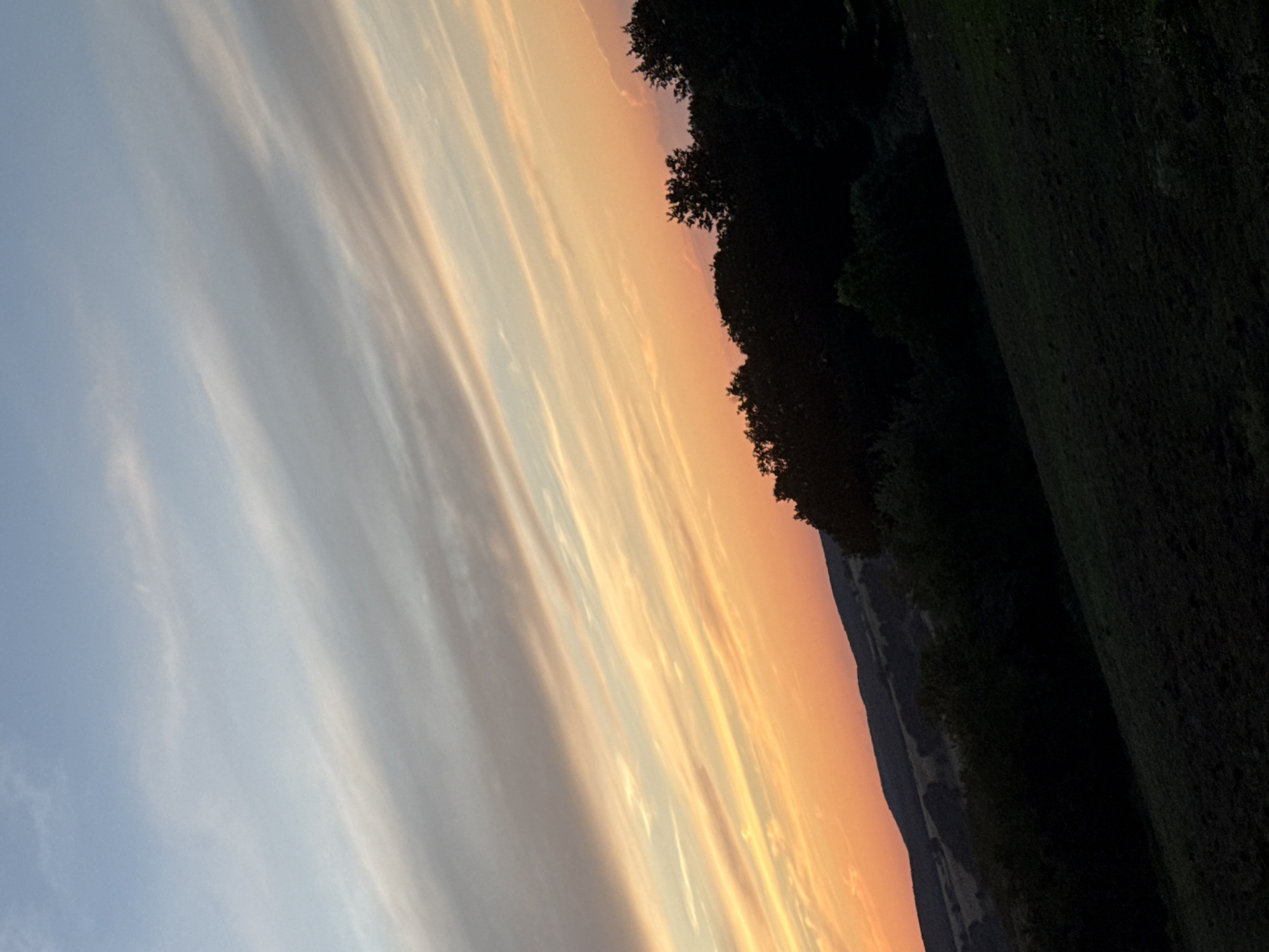 A wide-angle, low-angle shot captures a sunset over rolling hills in Krushevets, Bulgaria. The sky is filled with wispy, horizontal clouds in shades of pale blue, grey, and orange, creating a streaky effect. Below the sky, silhouetted trees and hills form a dark, undulating line across the frame. The foreground features a dark, sloped field. No people or animals are visible in the image. The overall impression is one of natural beauty and tranquility during twilight.