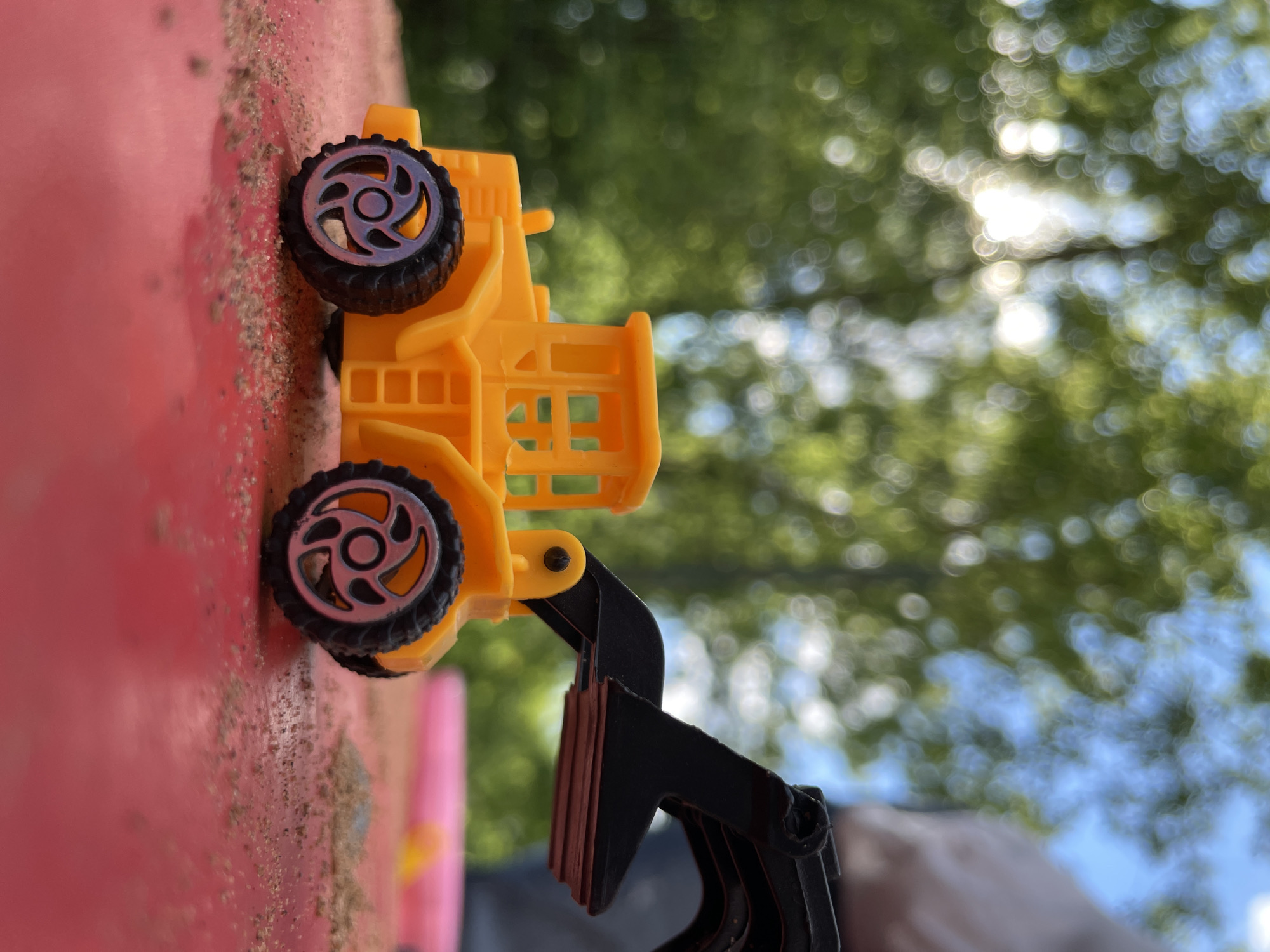 A close-up, sideways-oriented photograph features a bright yellow toy construction vehicle, possibly a front-end loader, set outdoors in Izhevsk, Russia. The toy is made of yellow plastic with black wheels that have greyish-silver multi-spoke hubcaps. Its black articulating arm and scoop, which has reddish-brown tips, are visible at the bottom right.
The toy rests on a reddish-pink surface, possibly a sandbox edge or a plastic play table, with scattered grains of sand or dirt. The background is heavily blurred with significant bokeh, showing vibrant green foliage and numerous bright, circular spots of sunlight filtering through leaves, indicating an outdoor, sunny daytime setting. Patches of light blue sky are also visible. No people or visible text are present in the image.