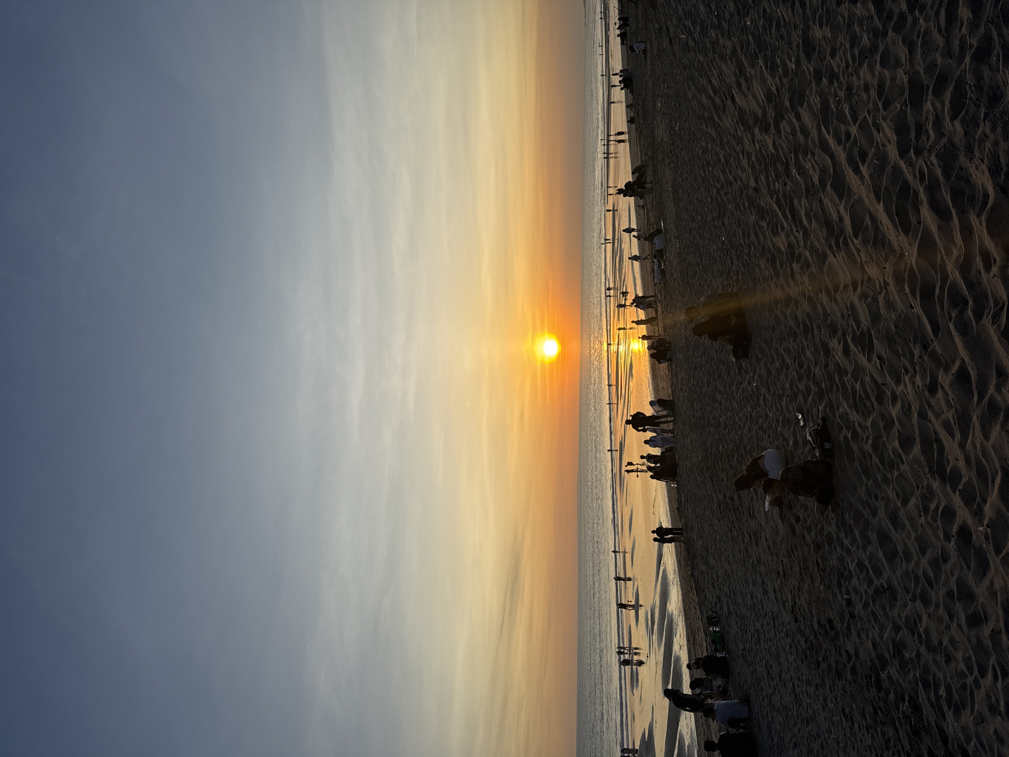 Captured at sunset in North Kuta, Indonesia, the image depicts a crowded beach. People are scattered along the shoreline, some walking, others sitting or standing near the water. The setting sun casts a golden glow over the sea and sky, creating reflections on the wet sand. The sky transitions from a light blue to a gradient of yellow and orange near the horizon, where the sun is setting. The beach is a dark color, contrasting with the bright sky and water. Footprints and ripples are visible in the sand.