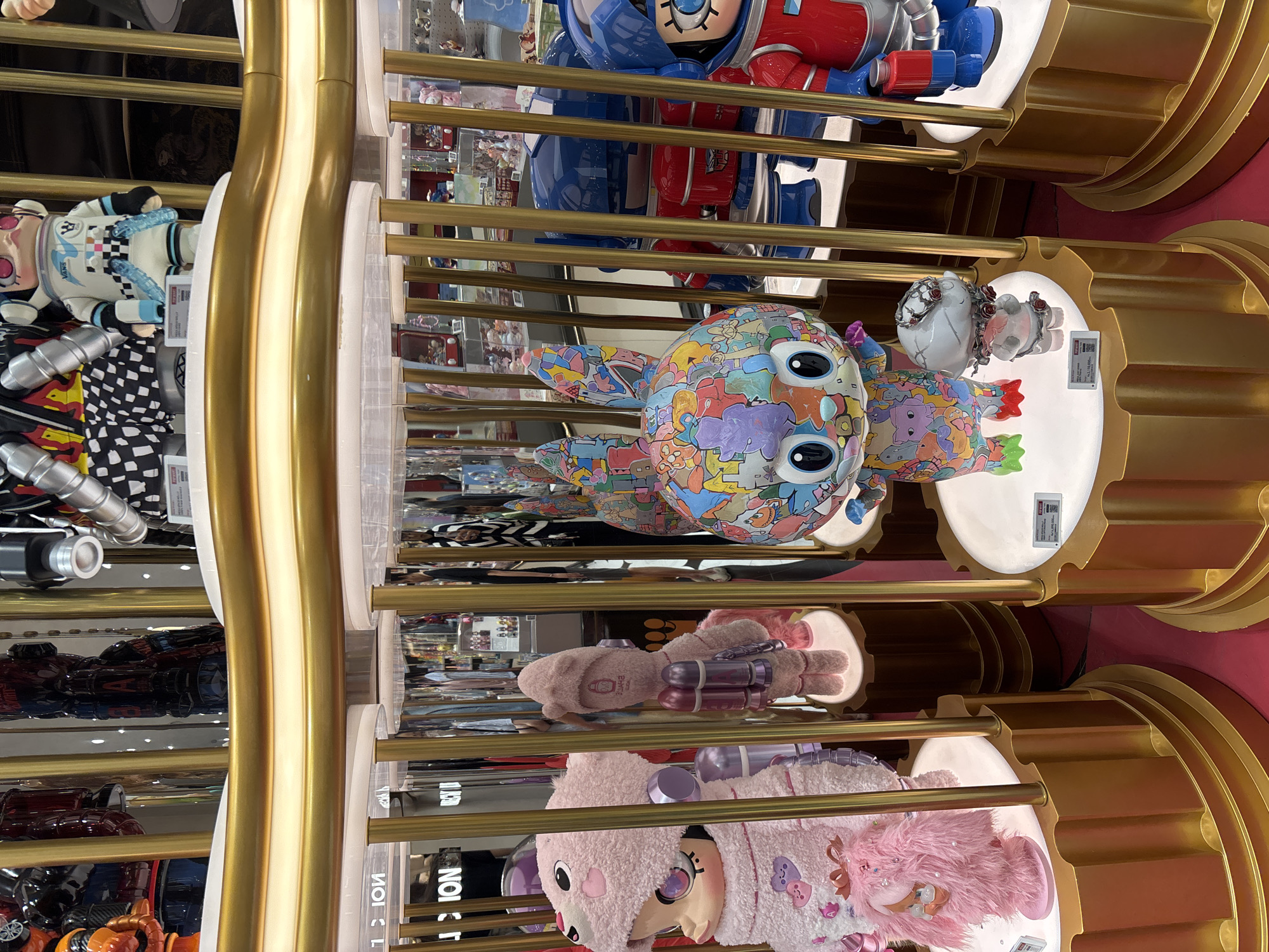 A display of toys behind a gold-colored metal frame is seen in an upright indoor shot. The toys include, from top to bottom, a red and blue astronaut-like figure, a bunny-shaped figurine with colorful designs on its body, a small white figurine, a light brown teddy bear-like figure, and a pink teddy bear-like figure. The toys are displayed on round white platforms with gold-colored cog-like bases. The image appears to have been taken in a shop in West Jakarta, Indonesia.