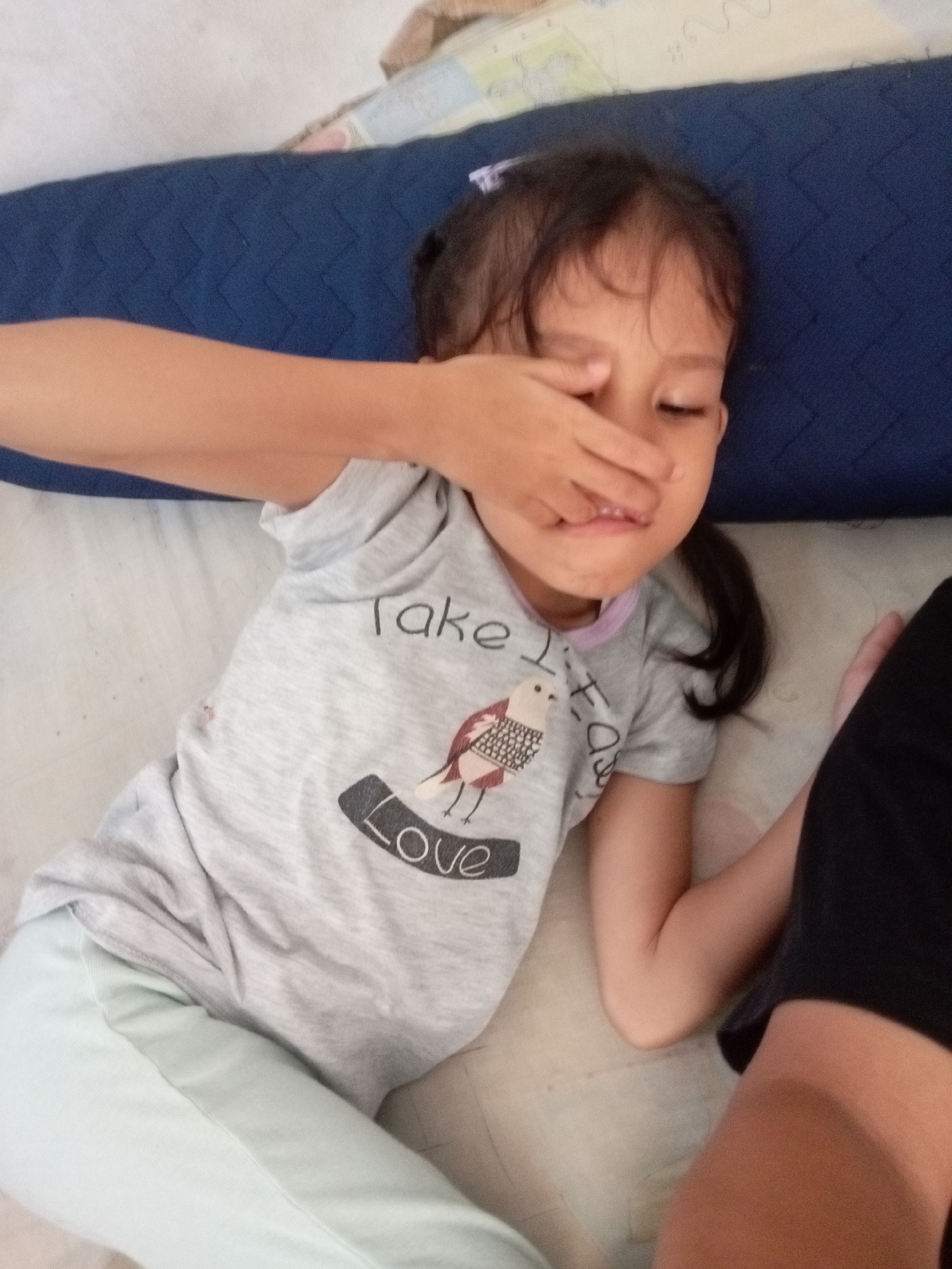 A young girl is lying down, resting her head on a dark blue pillow, with her right hand covering her right eye. She is wearing a light grey t-shirt with a graphic design and light blue sweatpants. The t-shirt has text that reads "Take love" and a drawing of a bird. The girl has dark hair pulled back in a ponytail and appears to be around 5-7 years old. The setting appears to be indoors, possibly a bedroom, with a light-colored bedsheet and a patterned blanket in the background. The lighting suggests it could be daytime, but the exact time of day is not discernible. The girl's expression is difficult to read due to her hand covering her eye, but she appears relaxed and possibly sleepy. There are no obvious weather cues. The image is likely taken in Batam, Indonesia, given the location context.