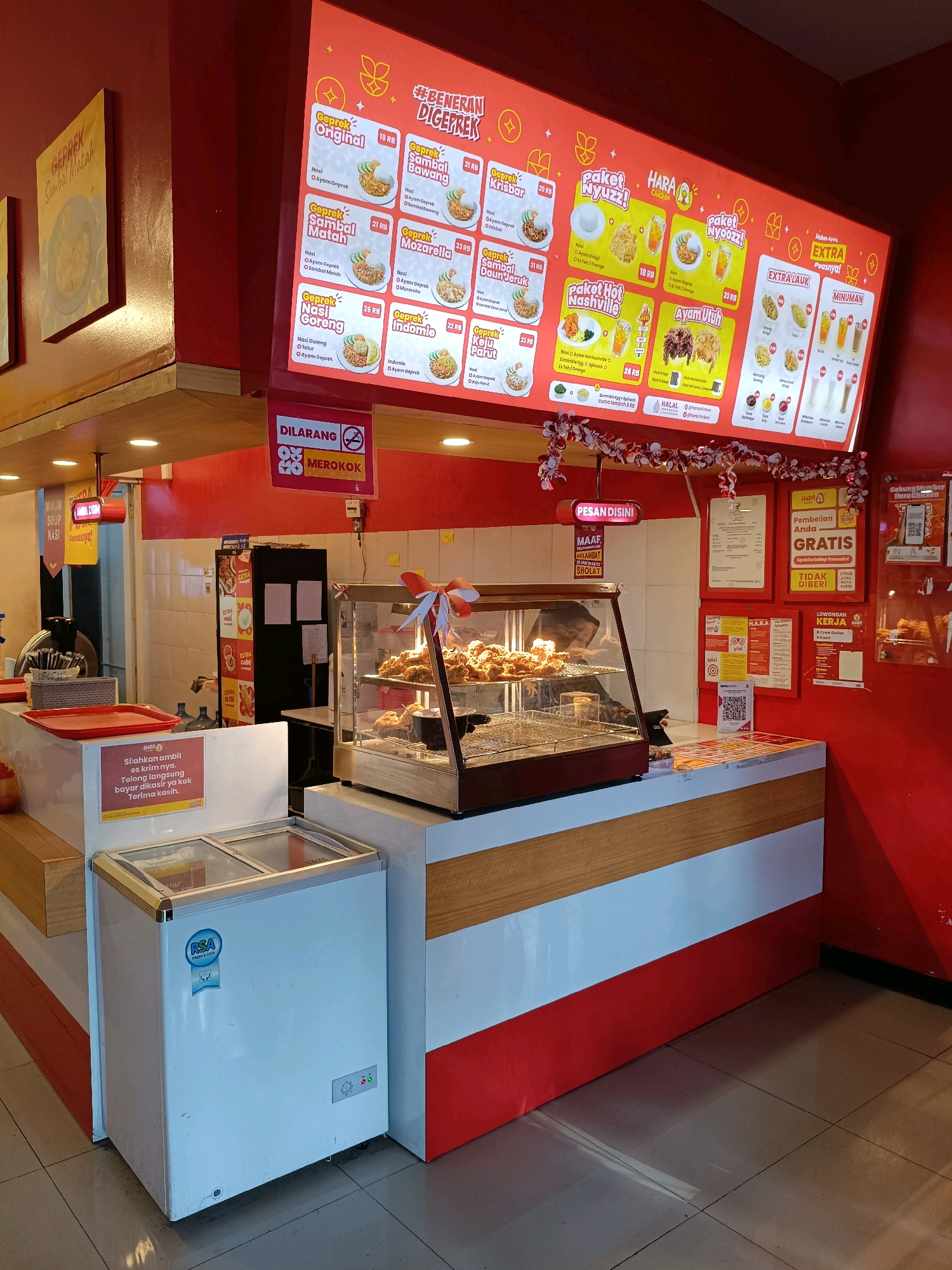 The image shows the interior of a fast-food restaurant, likely a fried chicken establishment, in Ngemplak, Indonesia. The restaurant features a prominent red and white color scheme with a large digital menu board displaying various chicken meals and combos with prices in Indonesian Rupiah (RB). Some menu items include "Geprek Original," "Geprek Sambal Bawang," "Geprek Krisbar," "Geprek Mozzarella," "Geprek Nasi Goreng," and "Geprek Indomie." There are also sections for "Paket Nyuzz!" and "Ayam Utuh," suggesting combo meals and whole chicken options.
In the foreground, a white chest freezer is visible. Behind it is a counter area with a glass display case showcasing fried chicken. To the right of the counter, a shelf displays drinks and condiments. Various signs are posted, including a "DILARANG MEROKOK" (No Smoking) sign, a "PESAN DISINI" (Order Here) sign, and information about promotions like "Pembelian Anda GRATIS" (Your Purchase is FREE). Another sign indicates "LOWONGAN KERJA" (Job Vacancy) for "Crew Outlest & Kasir."
The environment appears clean and functional, with tiled floors. The overall impression is that of a typical quick-service restaurant. There are no visible customers or staff actively engaged in transactions in this particular frame, though the setup implies an operational business. The decor includes festive red and white streamers.