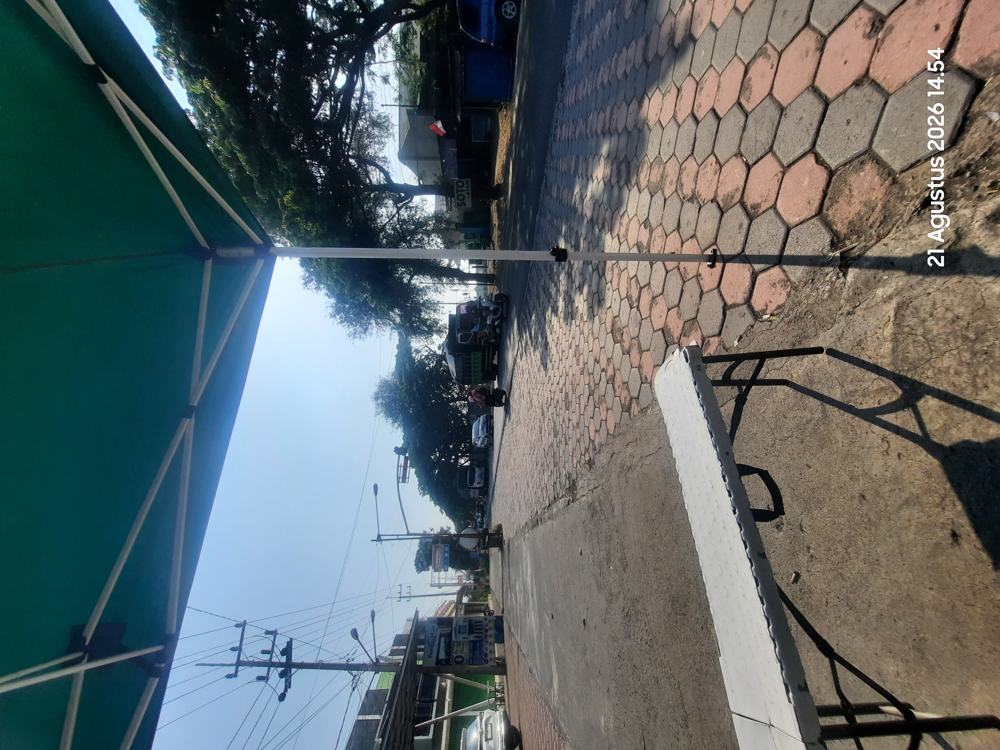 A low-angle shot, taken from beneath a green canopy, reveals a street scene in an urban environment. The foreground features a white folding table with black legs, casting a distinct shadow on the concrete and hexagonal paver ground. A white pole, supporting the canopy, stands to the right of the table. Beyond the table, a paved road stretches into the distance, lined with buildings on the left and trees on the right. Traffic is visible on the road, including a truck, a motorcycle with a rider, and other vehicles. A utility pole with numerous wires stands on the left side of the road. The sky is clear and blue, suggesting a sunny day. The bottom right corner of the image displays a timestamp: "21 Agustus 2026 14.54".