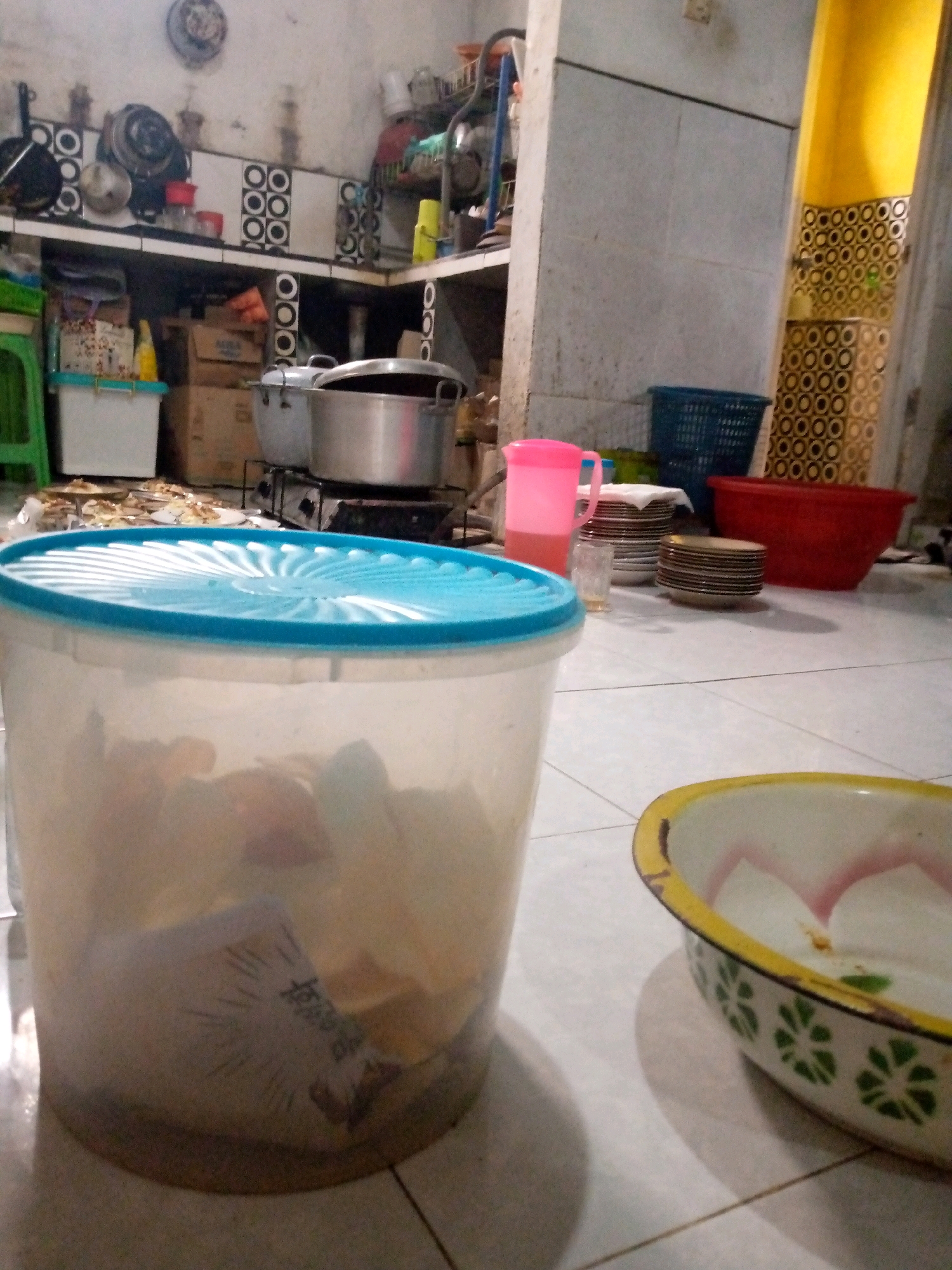 The image depicts a cluttered kitchen environment, likely in a residential setting in Sedati, Indonesia. The scene is filled with various kitchenware and household items. In the foreground, a large, transparent plastic container with a blue lid is prominent. Inside, what appears to be food items and some crumpled paper are visible. To the right, a small, decorative bowl with a yellow rim and floral green pattern sits on the tiled floor.
In the mid-ground, a stack of plates is seen, some covered with a white cloth. Adjacent to the plates is a tall, pink plastic pitcher containing a pink liquid, suggesting it holds a beverage. A blue basket, likely used for laundry or storage, is placed behind the plates. Further back, a large red bowl is visible. The kitchen also features cooking equipment, including pots and a stove, suggesting ongoing or recent food preparation. Shelves are laden with more containers, boxes, and cooking utensils. The walls are tiled with a black and white circular pattern in some areas, and the floor is tiled in a light color. The overall impression is one of a lived-in, functional kitchen, possibly reflecting the common household arrangements in Sedati, Indonesia.