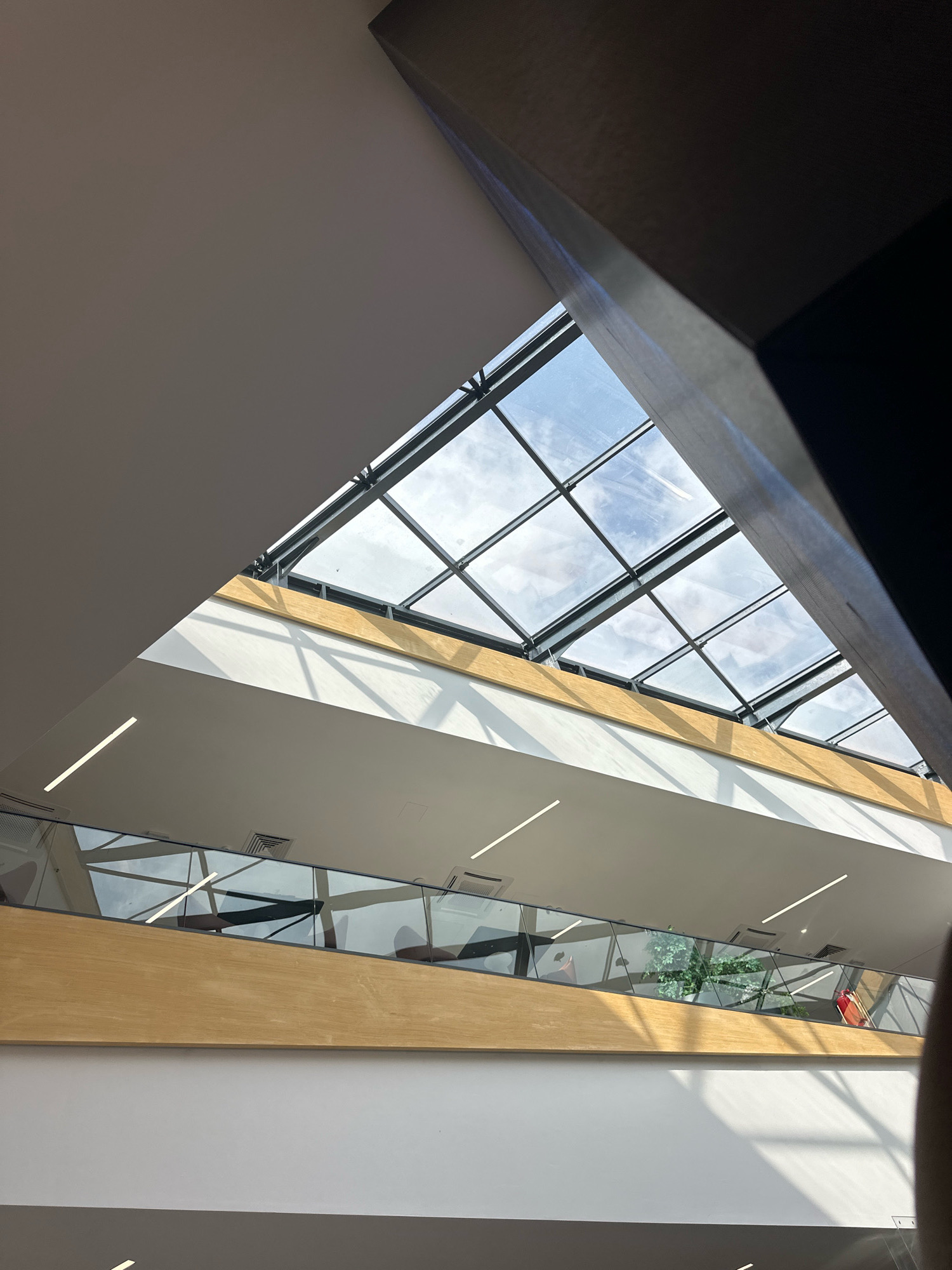 The image shows an interior view of a modern building with a large, angled skylight. The skylight is composed of multiple glass panes framed by dark metal beams, allowing natural light to enter the space. Below the skylight, there are multiple levels of walkways or balconies with glass railings. These levels are illuminated by linear strip lights. A wooden accent runs horizontally along one of the upper levels, adding a natural element to the design. Shadows from the skylight's structure are cast across the interior surfaces, indicating a bright, sunny day. The environment appears to be a public space, possibly a shopping mall, airport terminal, or a large office building. There are no identifiable people or specific landmarks that would place this image in Budva, Montenegro.