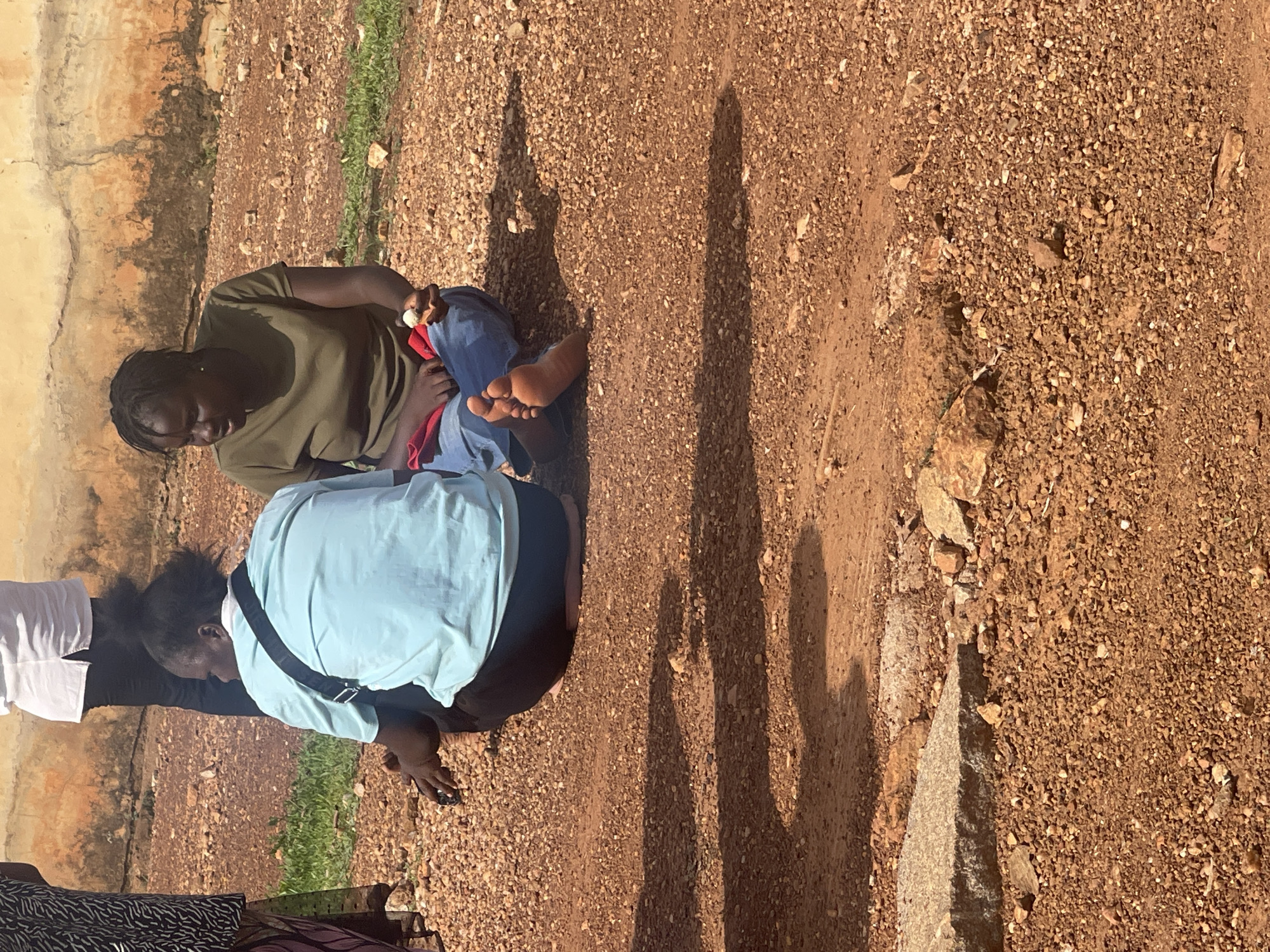 Two women are kneeling on a reddish-brown, gravelly ground surface. The woman on the left is wearing a light blue shirt and a black bag is slung across her shoulder. The other woman is wearing an olive-green shirt and blue pants. The location is Akwanga, Nigeria. A wall is visible to the left, and shadows are cast on the ground surface.