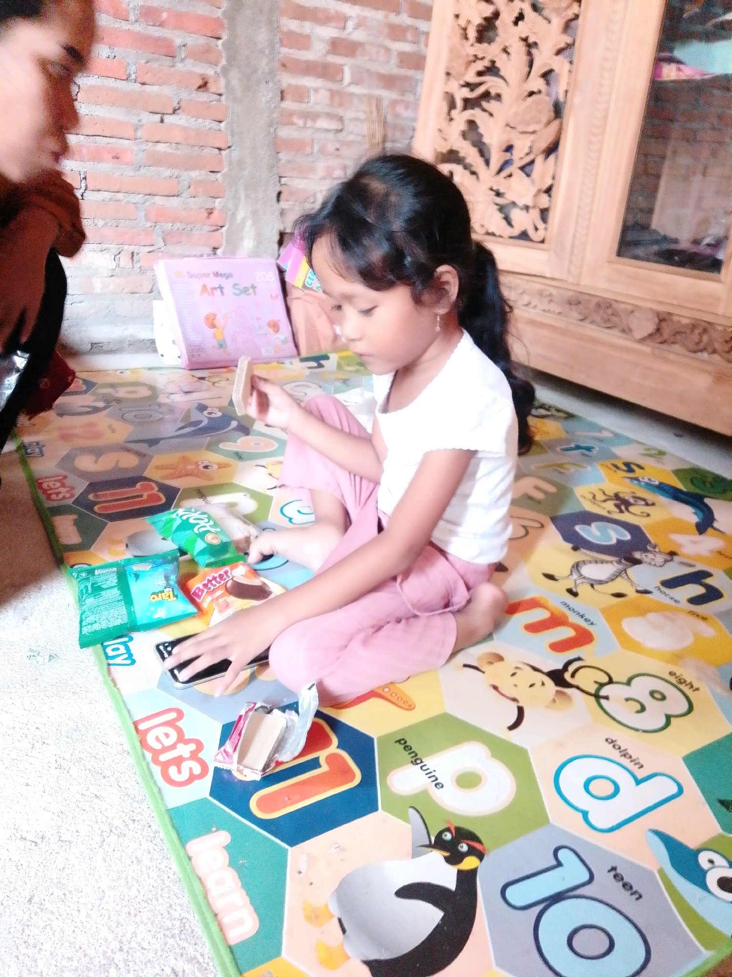 A young girl with dark hair tied in a ponytail, wearing a white t-shirt and pink pants, is sitting on a colorful playmat. She is looking down, focused on a small, open packet of snacks and a smartphone. The playmat features hexagonal sections with letters, numbers, and cartoon images of animals. Scattered around her on the mat are several snack packets, including one green packet with white text and another blue packet with white text. A small book titled "Super Mega Art Set" is visible behind her. To the left, a person's shoulder and arm, wearing a dark sleeve, are visible in the foreground, out of focus. The background consists of a brick wall and a wooden cabinet with carved details. The scene appears to be indoors, likely in a residential setting in Cikal, Indonesia.
