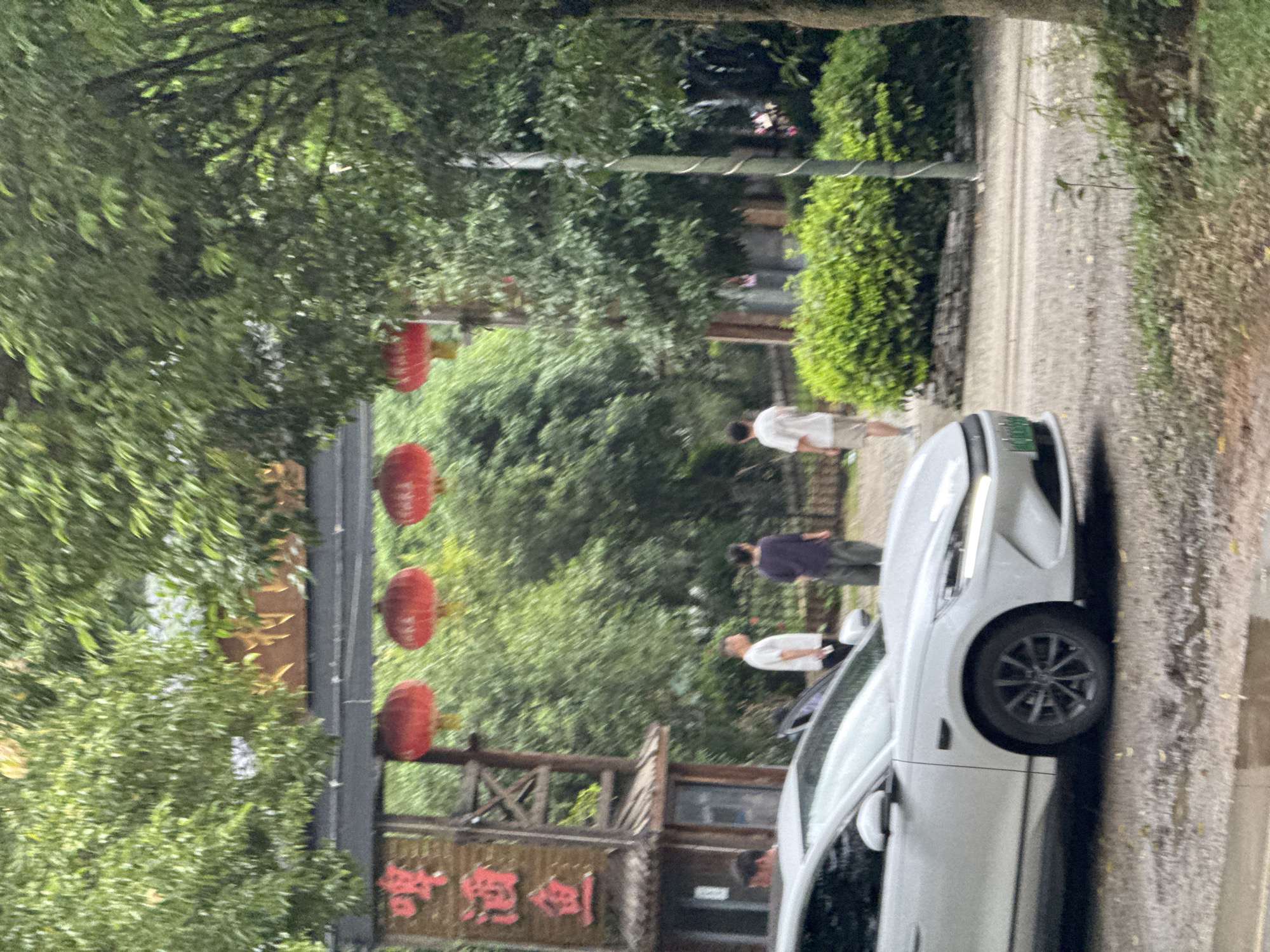 The image depicts a street scene in Guilin, China. Red lanterns are suspended above a wooden structure bearing Chinese characters. A white car is parked on a narrow road, beside a stone wall. Several people are walking along the road, away from the camera. Trees line the roadside, and a bush is also visible. The overall atmosphere is somewhat overcast.