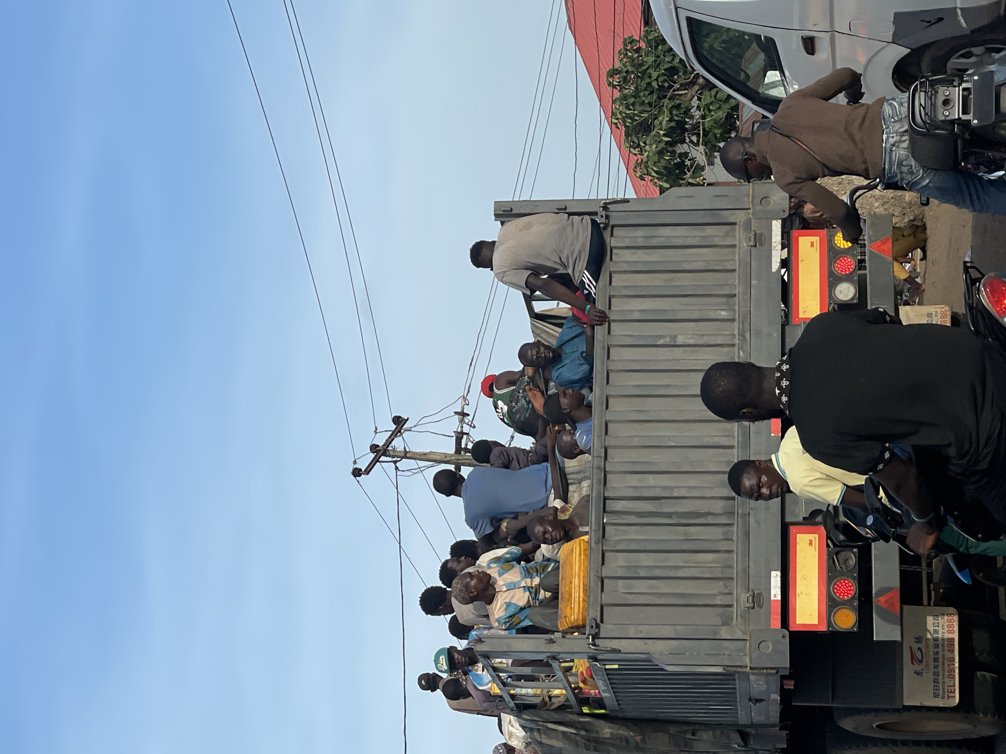 The image shows a truck filled with people, likely taken in Lokoja, Nigeria. The truck bed is crowded with individuals of various ages and attire. An overhead power line is visible above the truck. To the right of the truck, people are on motorcycles. The truck's rear has visible taillights and markings in Chinese characters along with the phone number 0916 498 8888 and the label "Nigeria longtemps industry co., ltd."