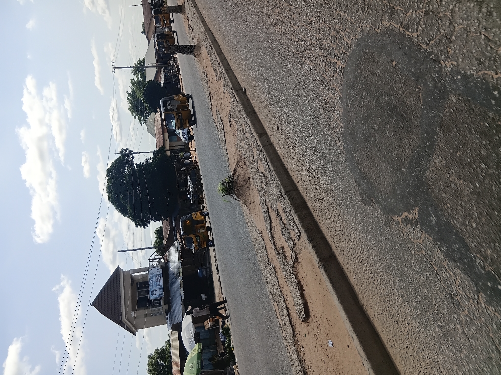 The image depicts a street scene in Jalingo, Nigeria, with a focus on a road and its surroundings. On the left side of the road, a building with a unique architectural design, featuring a prominent turret-like structure with a brown, conical roof, is visible. This building has windows and a balcony, and a banner is displayed on its facade. Adjacent to this building, there are stalls or shops with umbrellas providing shade, and some people are seen interacting in this area.
The road itself appears to be asphalt and shows signs of wear and tear, with several patches of damaged surface and a distinct darker patch on the right side, possibly indicating a repair or a different material. A concrete curb separates the road from a sandy embankment.
Several yellow auto-rickshaws, commonly known as Keke Napep, are present on the road. One is moving forward in the center of the frame, and at least three others are visible further down the road in a procession. A white car is also parked or moving on the left side of the road.
The background reveals a mix of buildings with corrugated iron roofs and trees, contributing to the urban and semi-urban atmosphere of the location. Overhead power lines stretch across the sky, which is partly cloudy. The overall impression is that of a typical street in a Nigerian city, with bustling activity and a blend of modern and traditional elements.