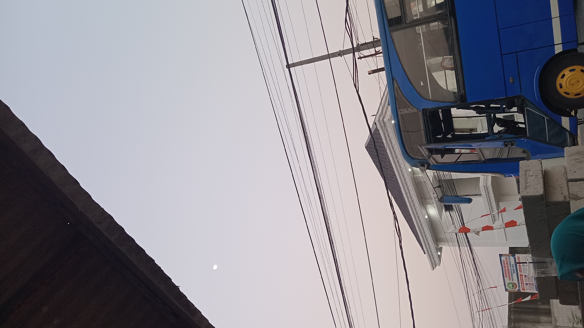 A blue bus is parked in front of a white building with red and white flags displayed. The building has a modern design with a dark roof and is adorned with lights. Several power lines crisscross the sky above the bus and building. In the distance, a faint moon is visible in the overcast sky. The foreground features a pile of concrete blocks, and a person wearing a teal head covering is partially visible, bent over in front of the blocks. A sign with Indonesian text is also present near the flags. The scene appears to be an outdoor setting, possibly in Trembulrejo, Indonesia, given the presence of the Indonesian flags and the visual cues of the environment.