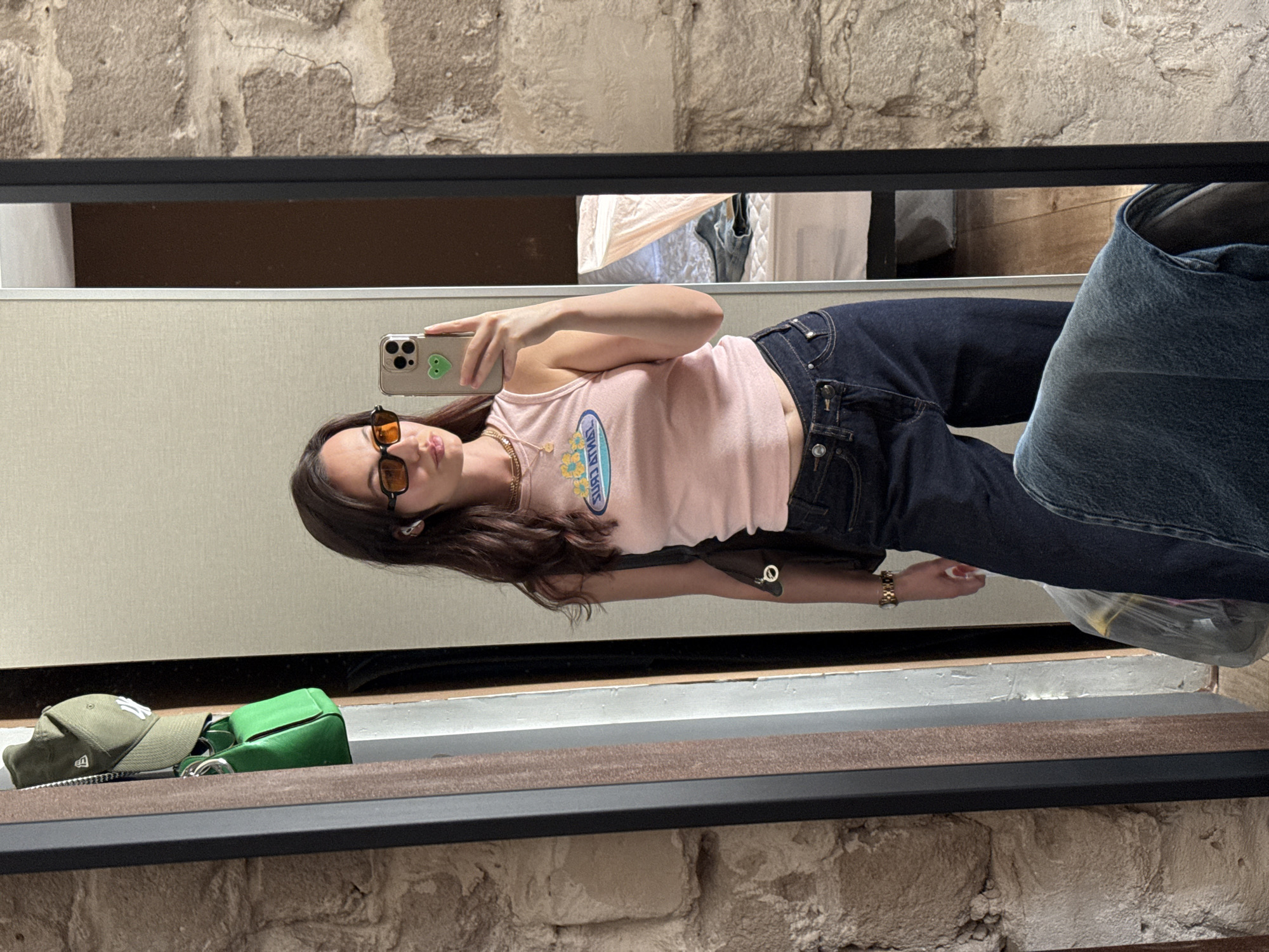 A young woman is taking a selfie in a mirror. She is wearing orange sunglasses, a pink tank top with a blue and yellow design, and dark blue jeans. The mirror is reflecting the room behind her, which appears to be a retail or changing room setting, including what may be a denim bag, a green purse, and a green baseball cap. The background includes a wall made of stone. While a location cannot be confirmed from the image alone, the likelihood of this being in Tel Aviv, Israel, is dependent on the source of the image.