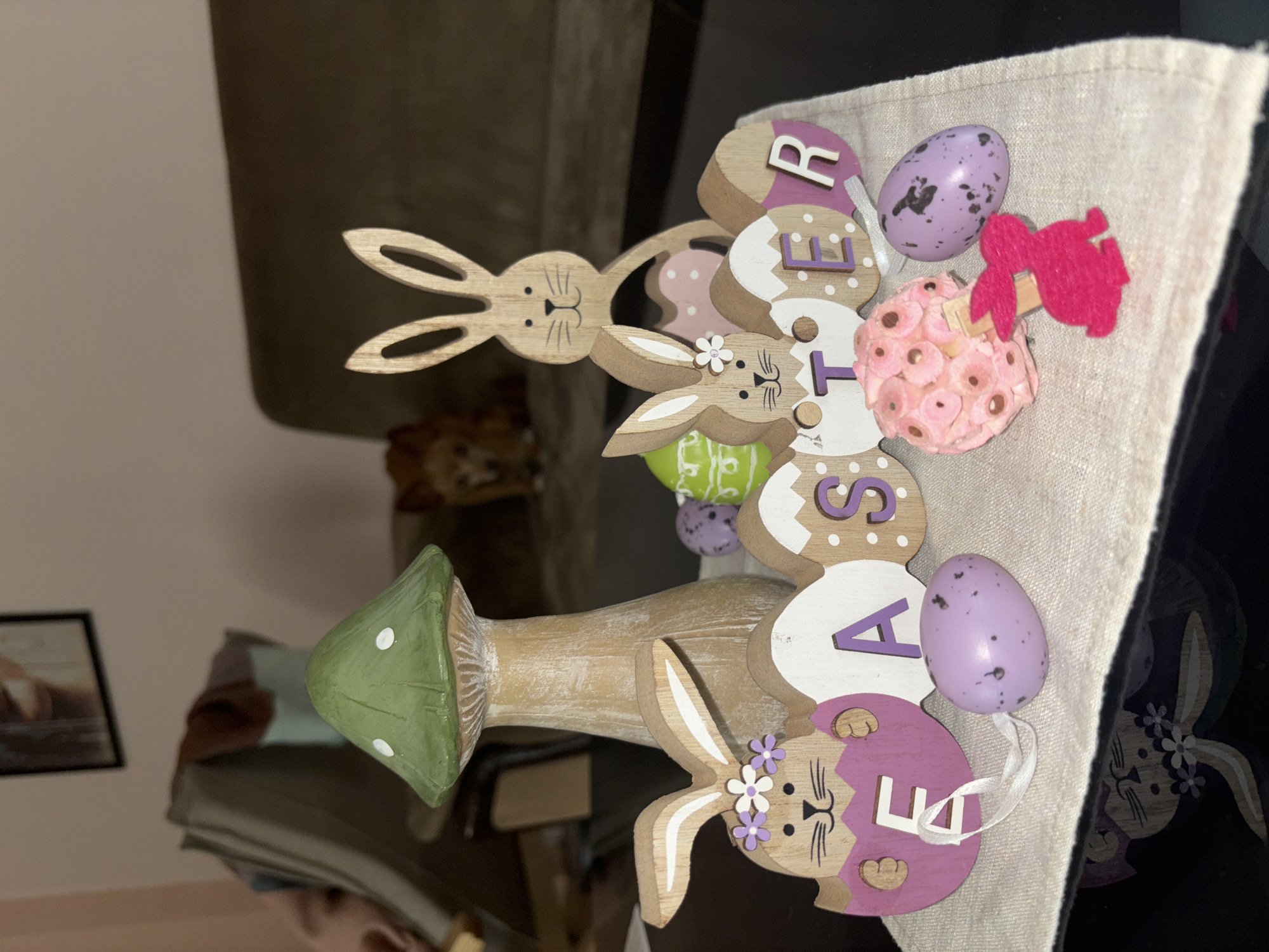 Here is a description of the image:
The image shows an Easter decoration resting on a light-colored surface, possibly a linen tablecloth. The decoration consists of a wooden mushroom base with a green cap, topped with a series of wooden discs stacked vertically to spell the word "EASTER". Between each disc is a small, painted Easter egg in colors such as pink, green, and purple. Three wooden rabbit figures are incorporated into the design, positioned between the discs. The location is not determinable.