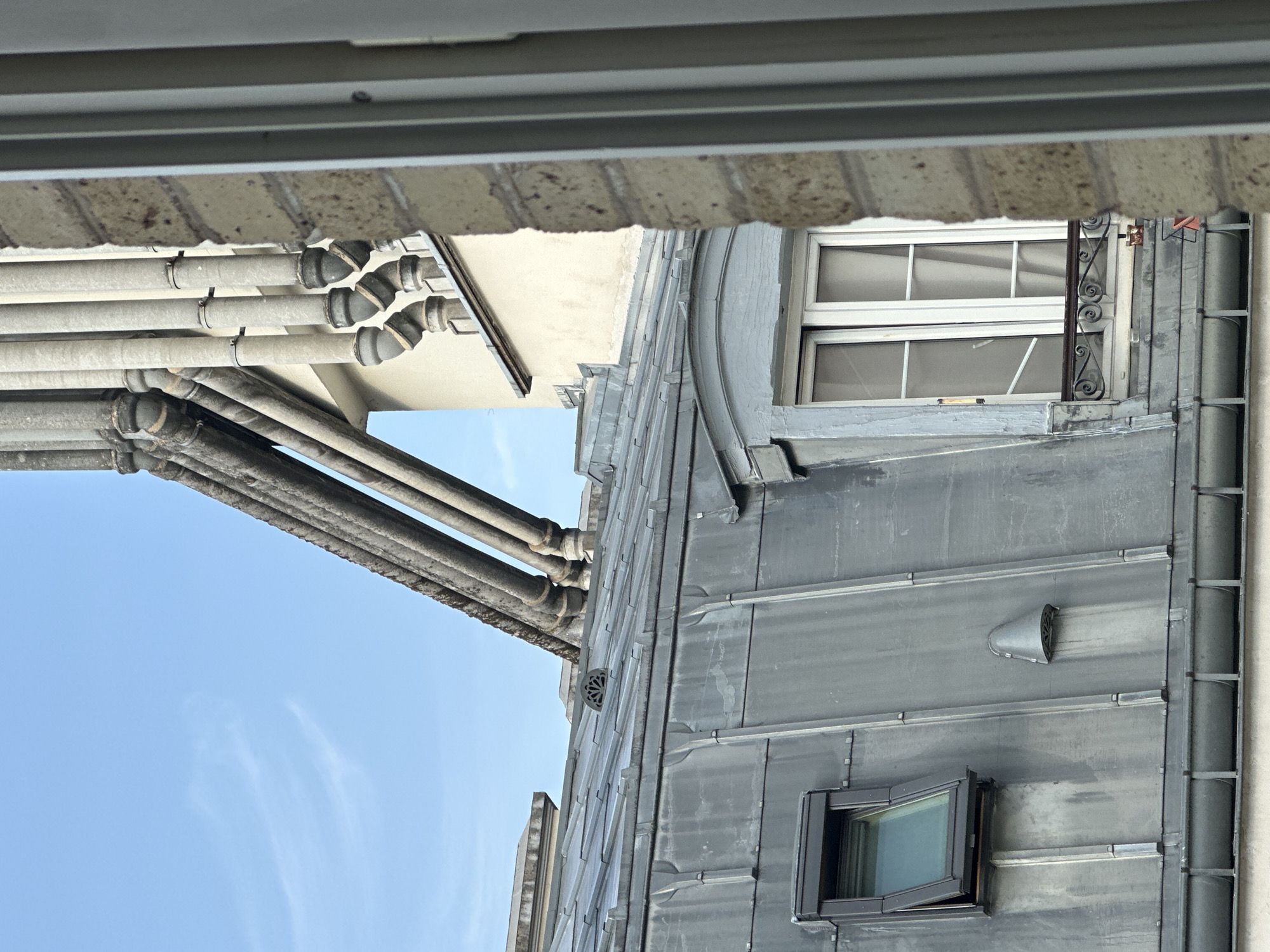 The image depicts a close-up view of a Parisian rooftop with a mansard roof made of grey metal sheeting. A skylight window, open at an angle, is visible on the left side of the roof. To the right, a dormer window with white sashes and curtains is set into a curved, grey frame. A decorative metal railing is present in front of the dormer window.
Above the mansard roof, a series of thick, grey pipes, likely ventilation or plumbing, ascend diagonally against a pale blue sky with faint wisps of clouds. These pipes are supported by ornate stone structures. In the background, a light-colored building with a dark cornice is visible, partially obscured by the pipes.
To the right edge of the frame, a portion of a brick wall with a textured, tan-colored surface is visible, along with a thin, vertical white frame, suggesting the perspective is from inside a building looking out through a window. The overall impression is one of architectural detail common in older Parisian buildings, particularly those with mansard roofs.